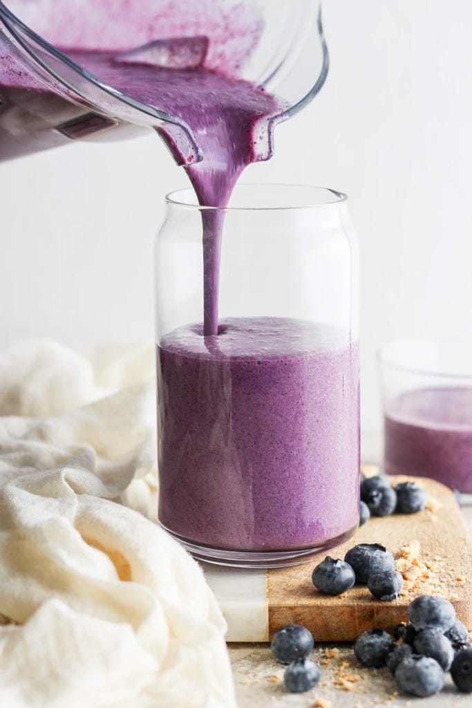 Pouring a fresh blueberry smoothie into a glass jar, surrounded by scattered blueberries on a wooden board.