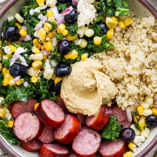 A bowl filled with quinoa, sliced sausage, kale, corn, black olives, onions, and a dollop of hummus.