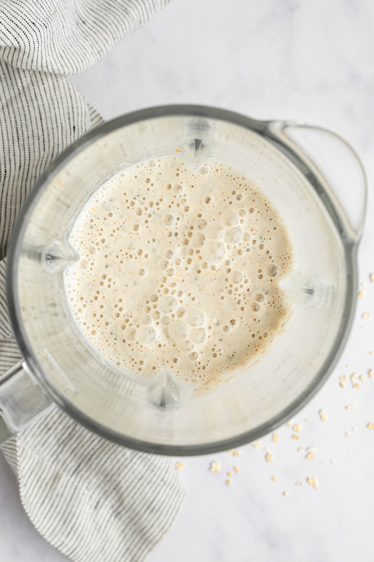 A blender filled with a frothy mixture is shown from above on a light-colored surface. A striped cloth and some spilled oats are visible next to it.