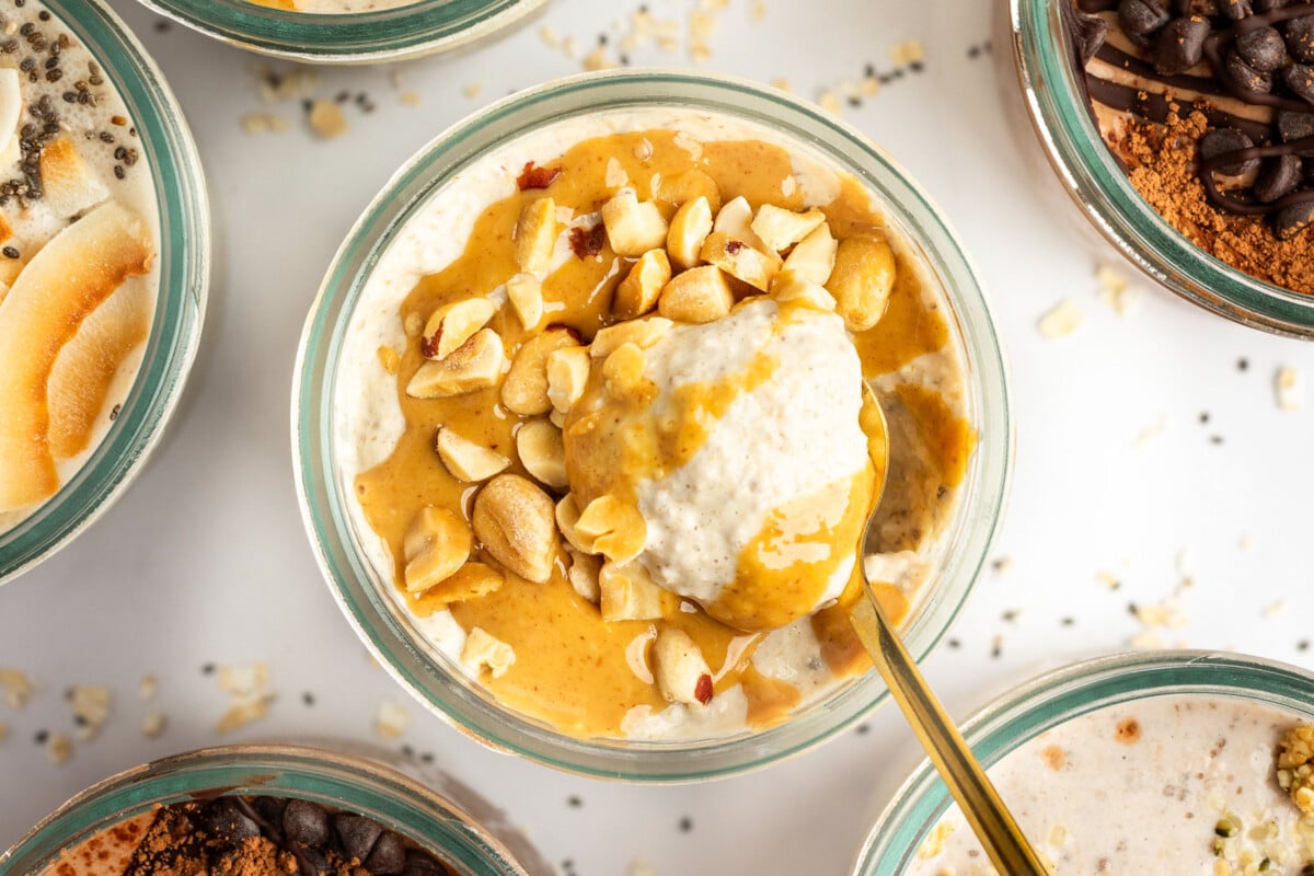 A bowl of creamy oatmeal topped with peanut butter and chopped nuts. A gold spoon is partially submerged in the oatmeal. Other bowls of similarly prepared oatmeal surround it.