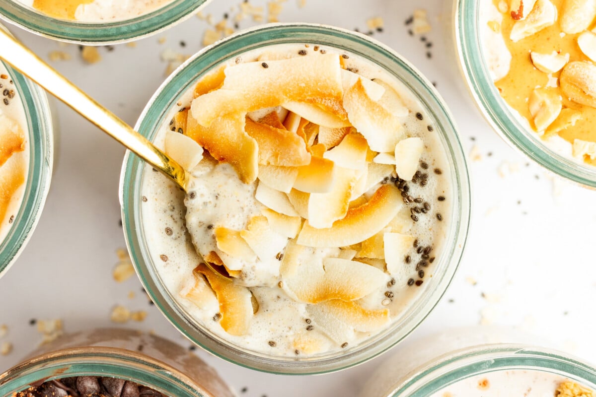 A glass bowl filled with a creamy mixture topped with toasted coconut flakes and chia seeds, with a gold spoon inserted into it. Other similar bowls are partially visible around it.