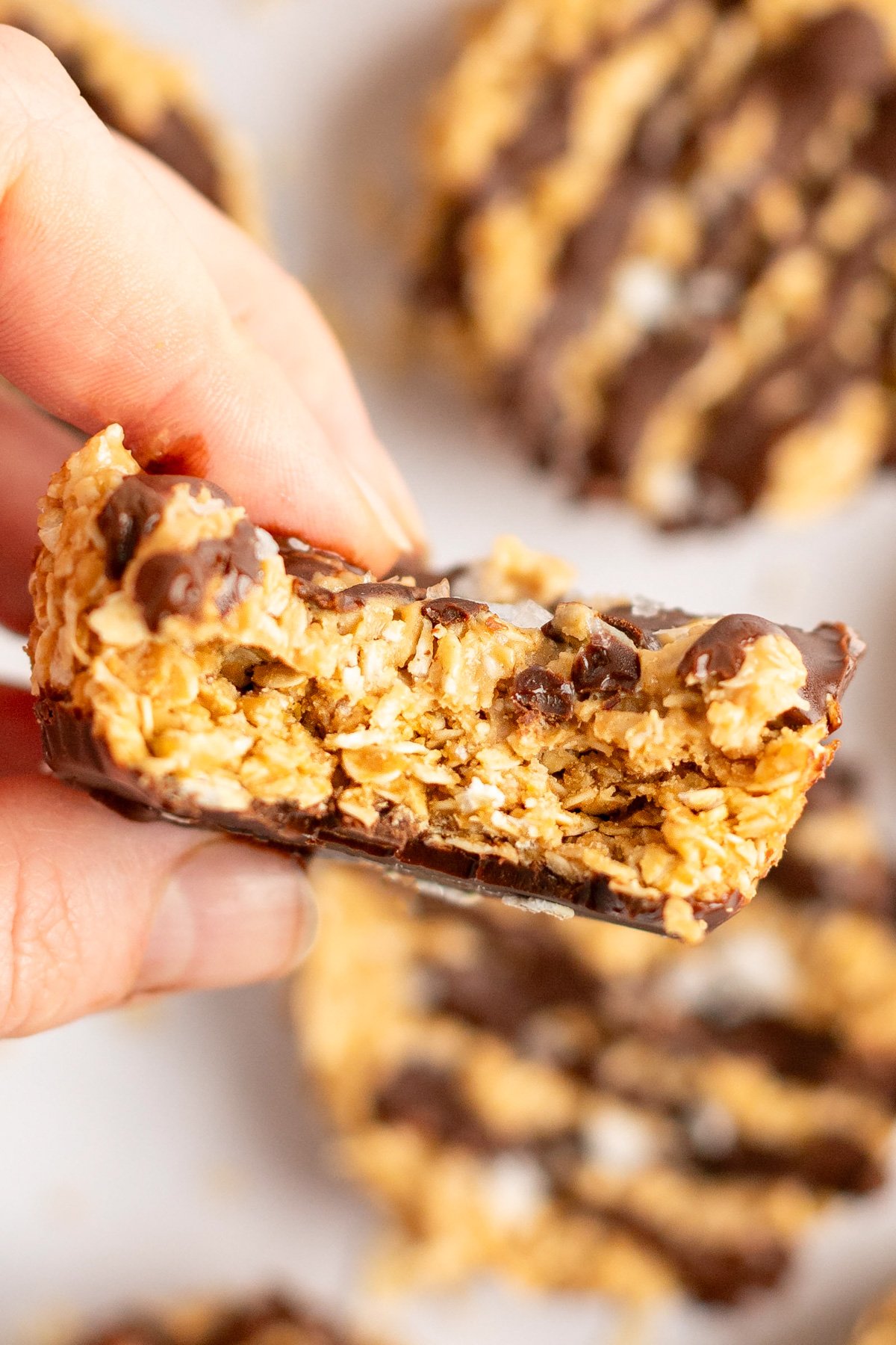 A hand holding a chocolate-drizzled oat bar with a bite taken out. Other similar bars are visible in the background.