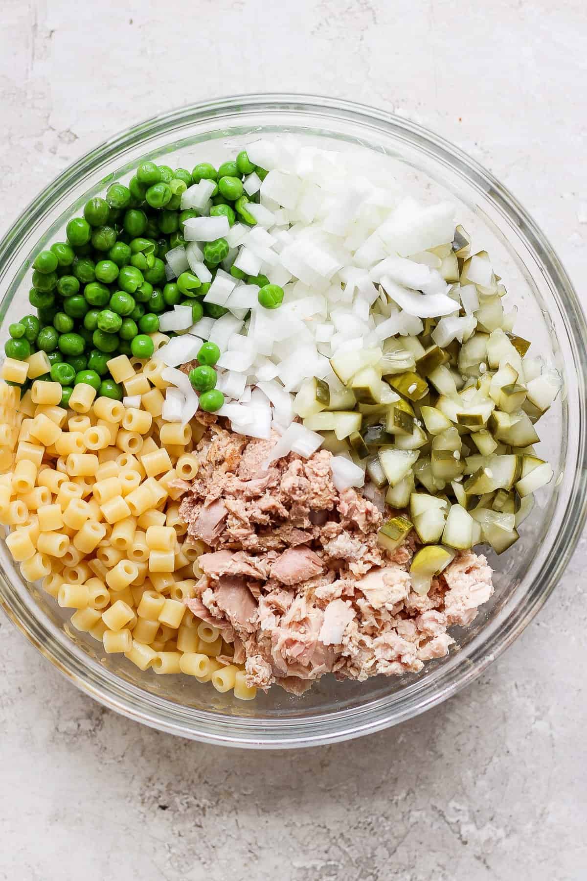 A bowl containing separate sections of diced onions, green peas, chopped pickles, macaroni pasta, and canned tuna, arranged on a light, textured surface.