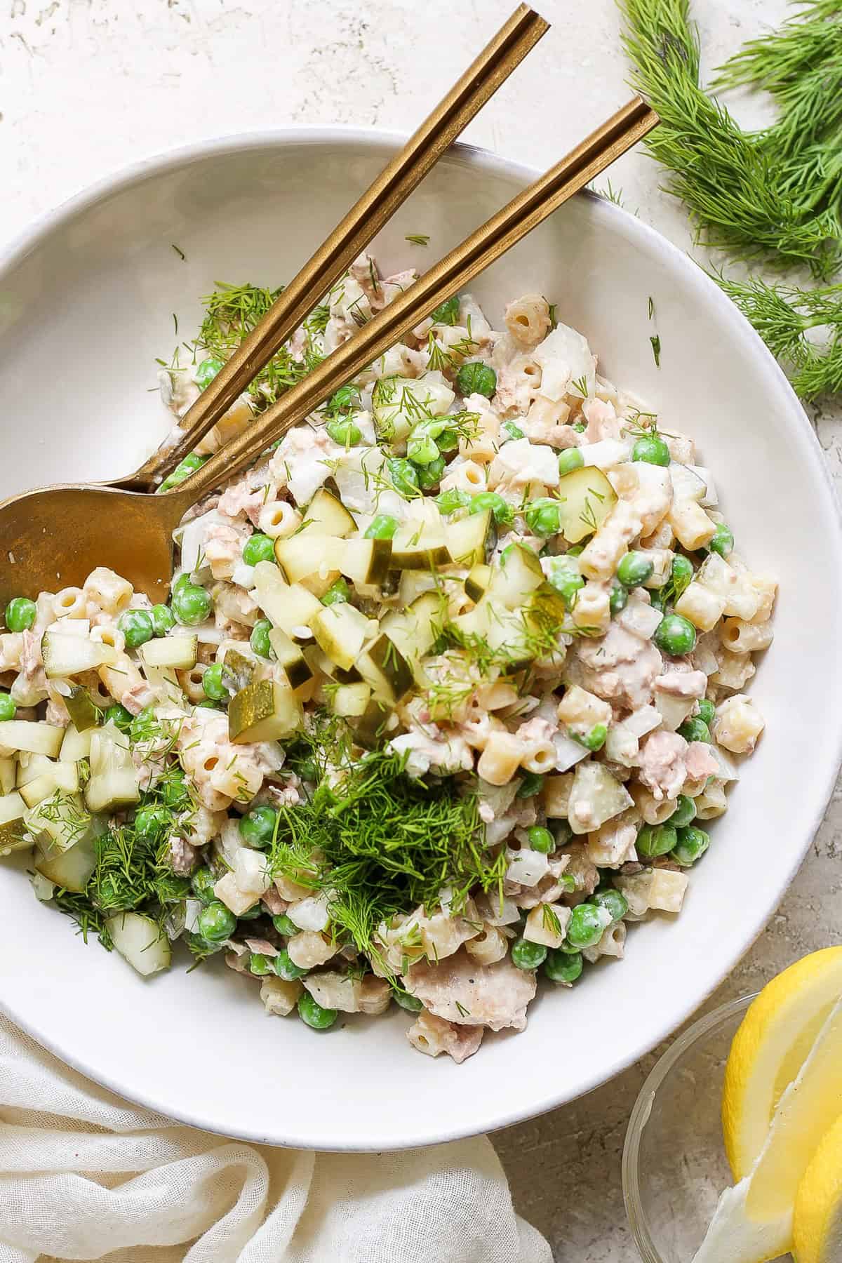 A bowl of tuna salad with peas, chopped cucumber, and dill, served with a pair of chopsticks and fresh lemon slices on a neutral-toned background.