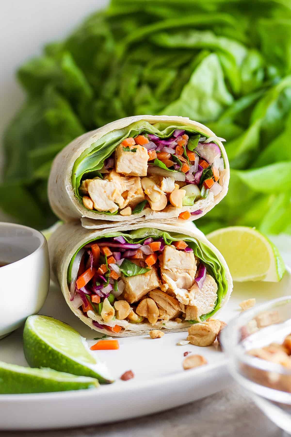 A chicken wrap with diced vegetables, peanuts, and lime wedges on a white plate, accompanied by a bowl of sauce; fresh lettuce in the background.