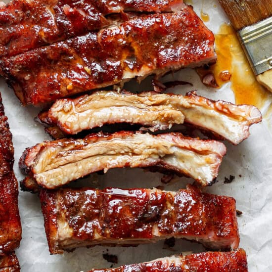 Close-up view of several pieces of glazed, barbecued ribs on parchment paper, with a brush coated in sauce to the side.