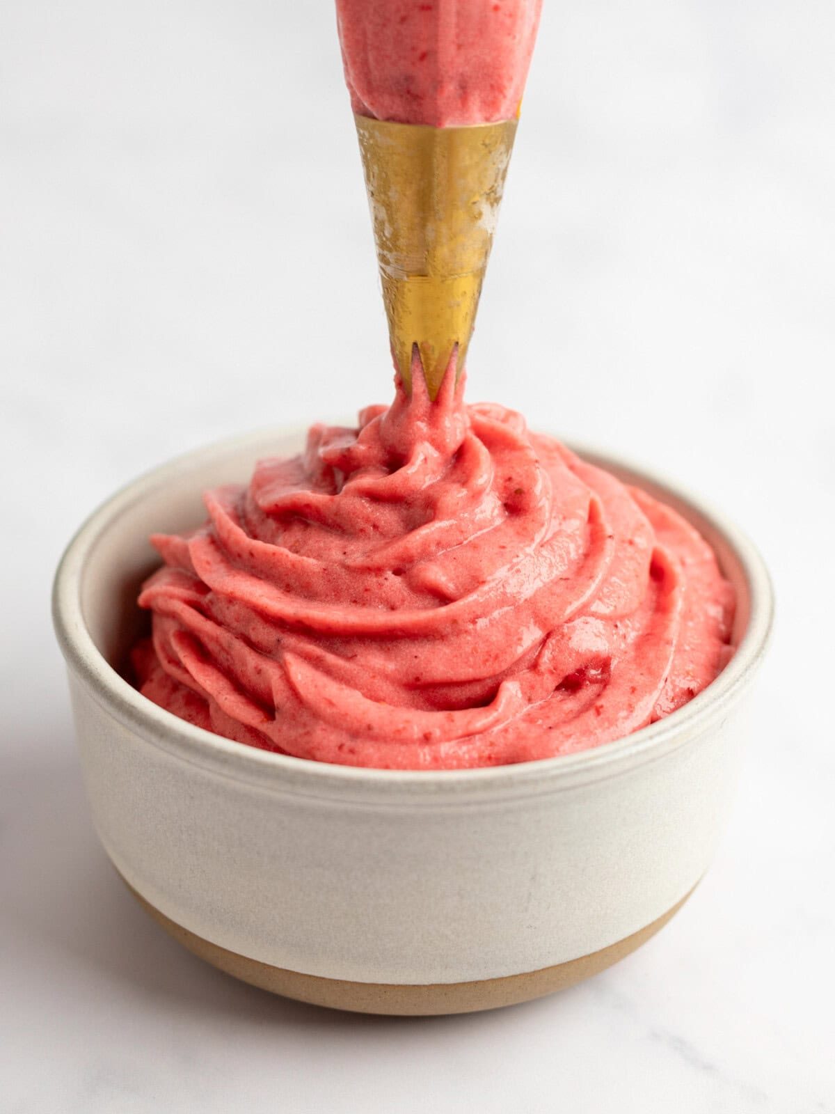 A piping bag dispensing creamy strawberry frosting into a small beige bowl against a white background.