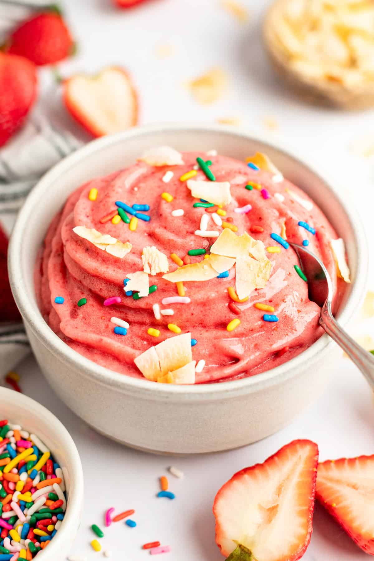 A bowl of pink strawberry banana smoothie bowl topped with sprinkles and coconut flakes, surrounded by fresh strawberries and sprinkles.