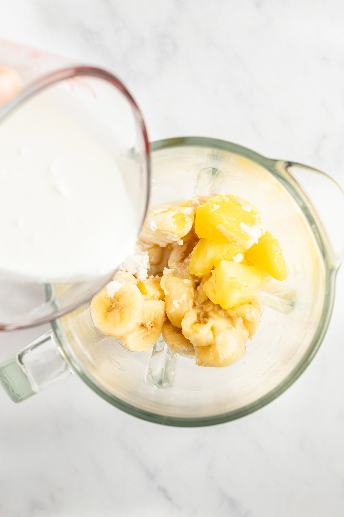 A measuring cup pours milk into a blender containing sliced bananas and pineapple chunks.