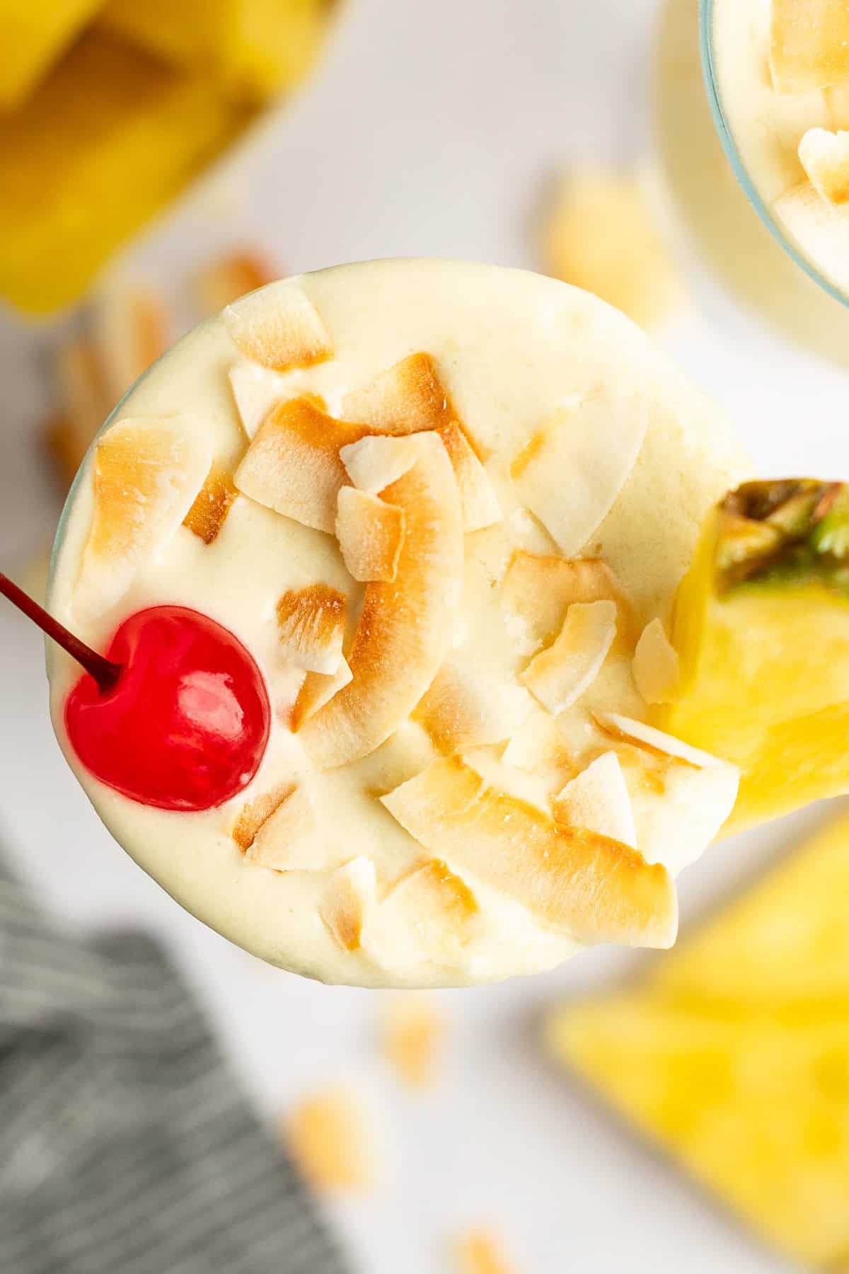 Top view of a creamy cocktail garnished with toasted coconut flakes, a maraschino cherry, and a pineapple wedge.