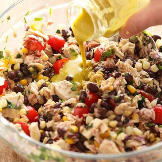 A hand pouring dressing into a glass bowl filled with a vibrant fiesta tuna salad, brimming with beans, corn, tomatoes, herbs, and chunks of tuna.