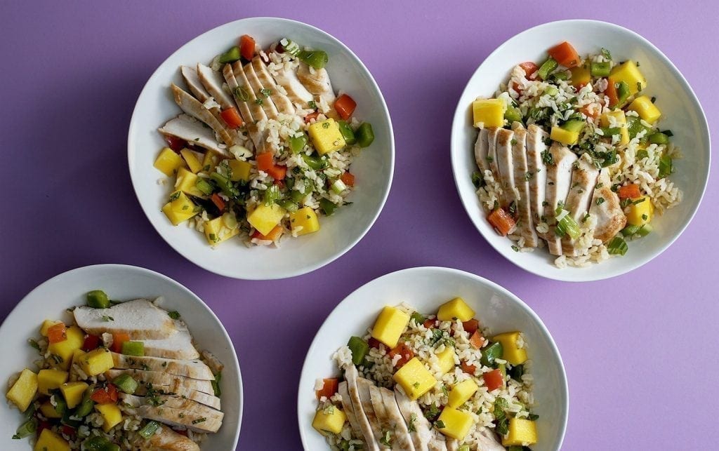 Four white bowls filled with sliced grilled chicken, diced mango, and a mix of bell peppers placed on a bed of rice. The bowls are arranged on a purple surface. MyFitnessPal Blog