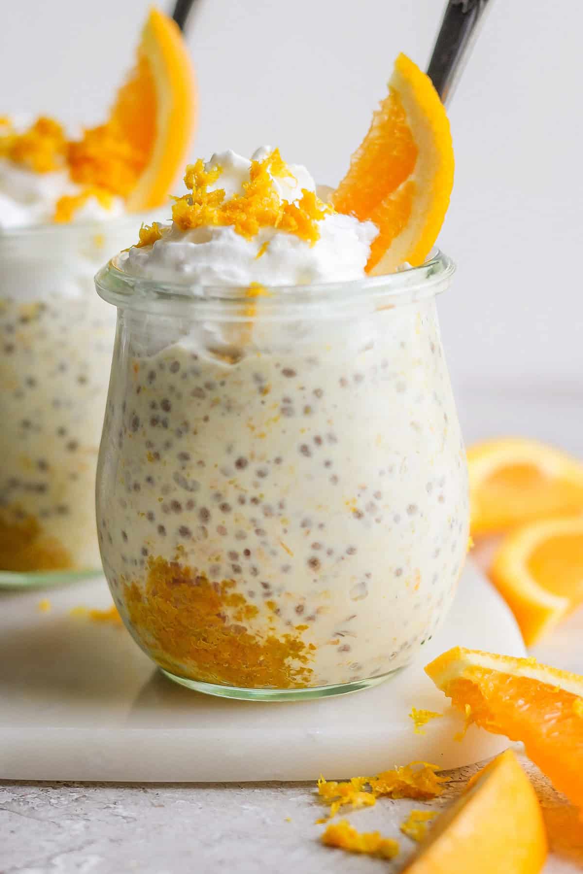 A glass jar filled with chia seed pudding topped with whipped cream, orange zest, and an orange slice, giving it a delightful creamsicle overnight oats feel, accompanied by pieces of orange on a white surface.