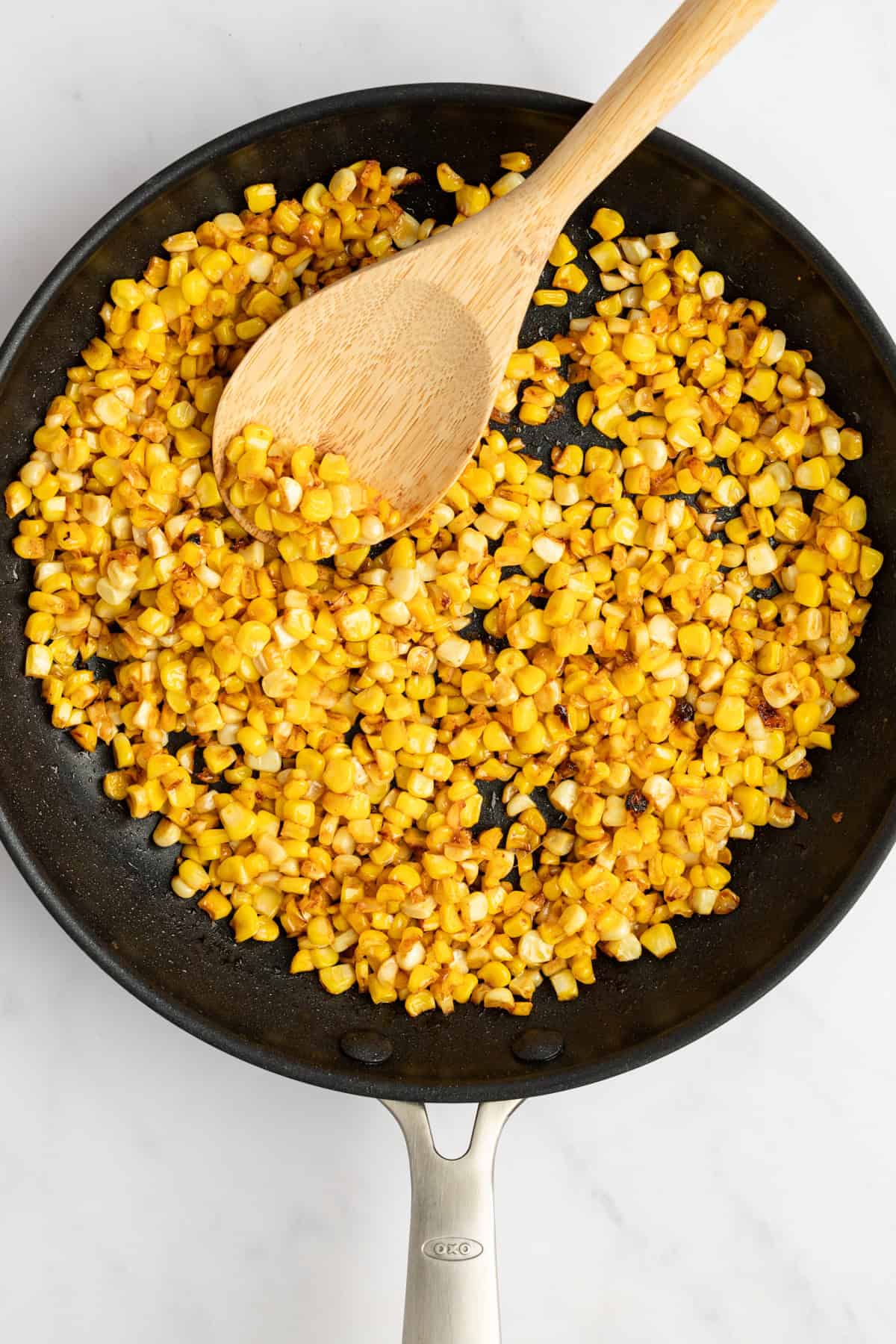 Sautéed corn kernels in a black frying pan with a wooden spoon on a white countertop.