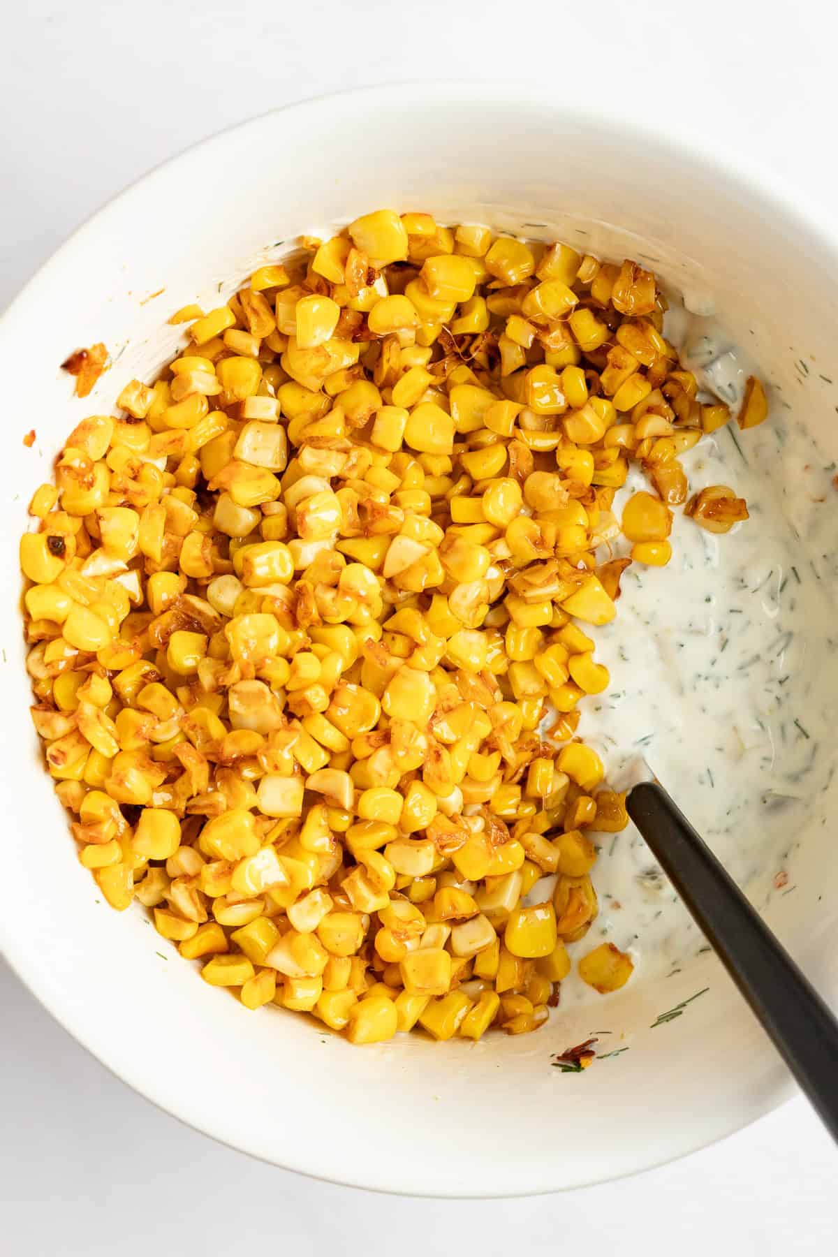 A bowl of roasted corn kernels mixed with a creamy, herb-infused dressing, and spoon partially immersed.