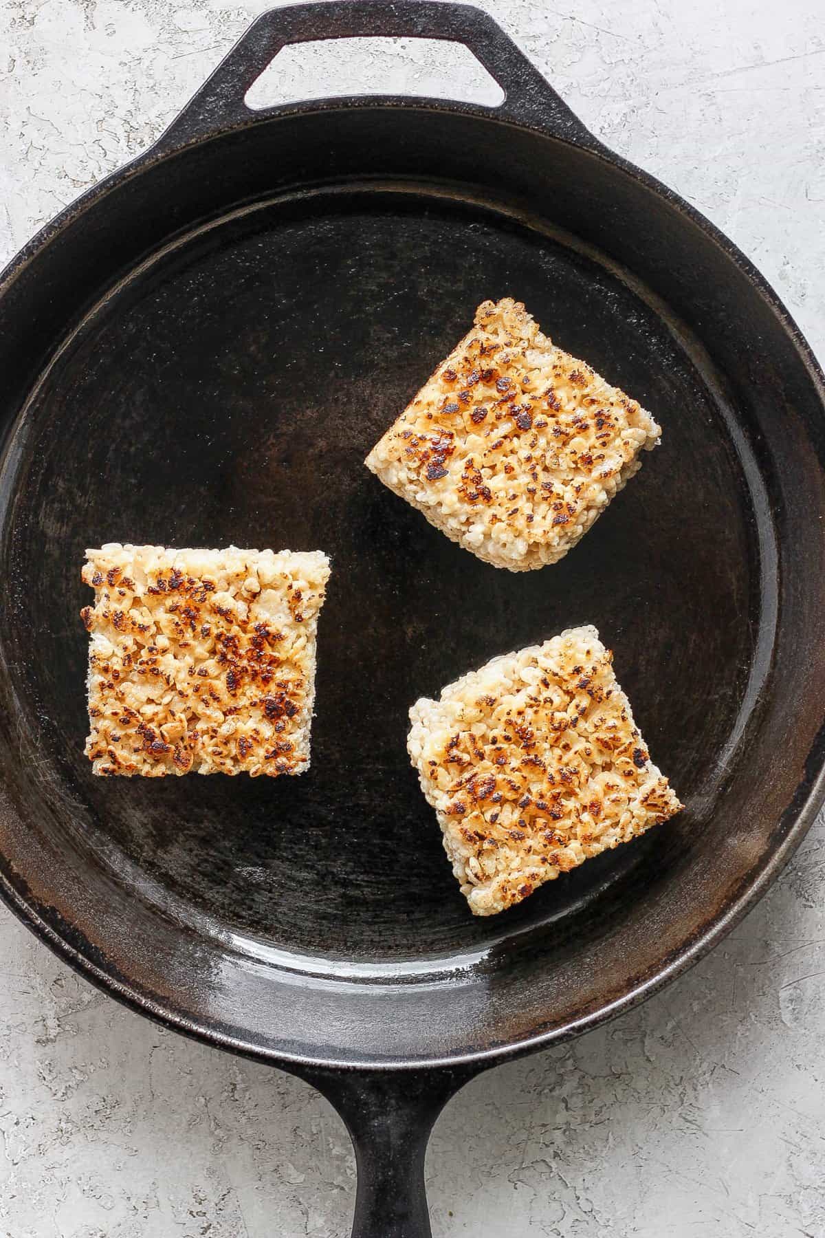 Three toasted Rice Krispie treats in a cast iron skillet on a light textured surface.