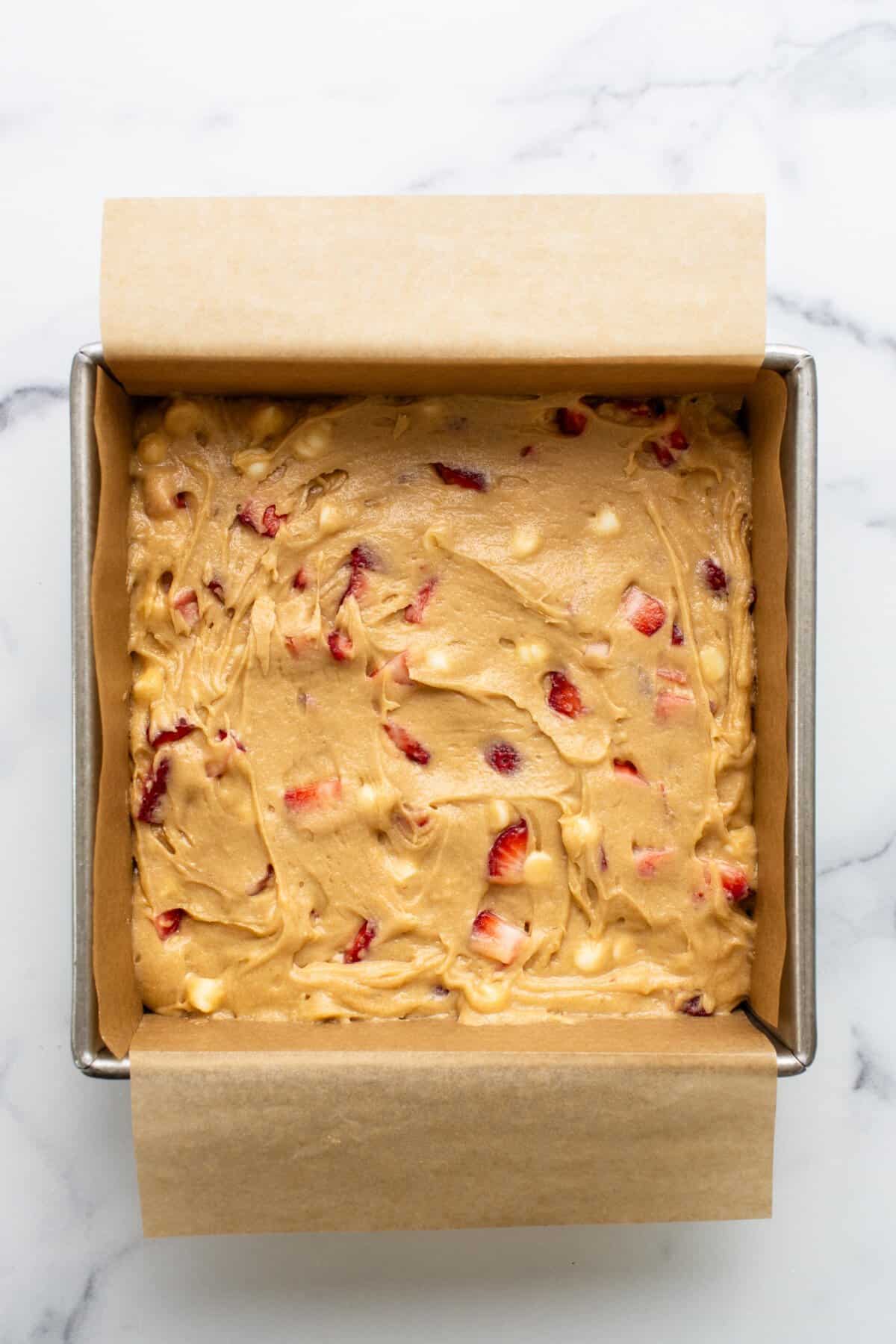 Square baking pan lined with parchment paper and filled with raw batter containing chunks of fruit, promising delicious strawberry blondies on a marble countertop.