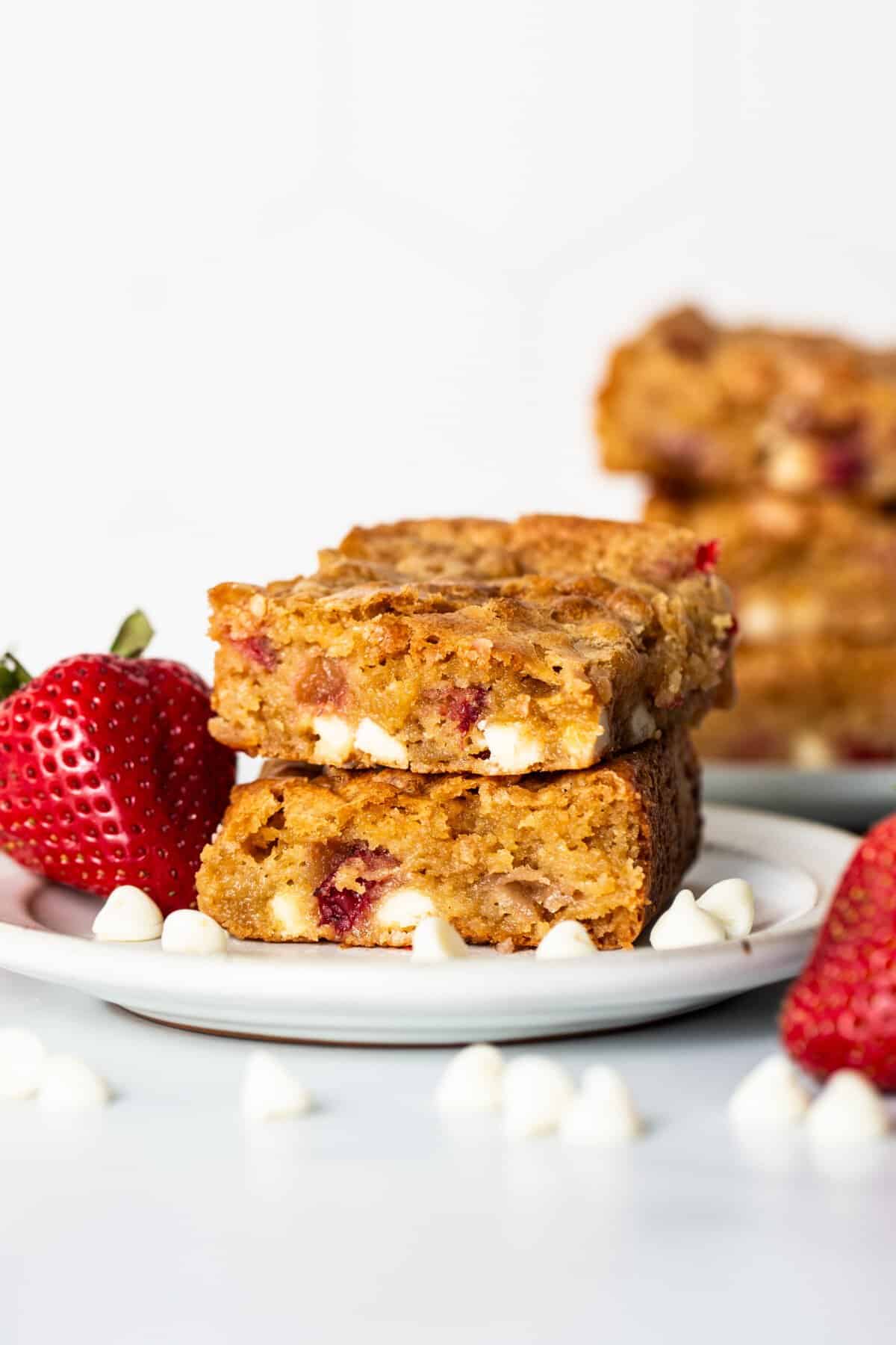 Close-up of two stacked strawberry blondies with visible strawberries and white chocolate chips on a small plate, accompanied by whole strawberries and scattered white chocolate chips.