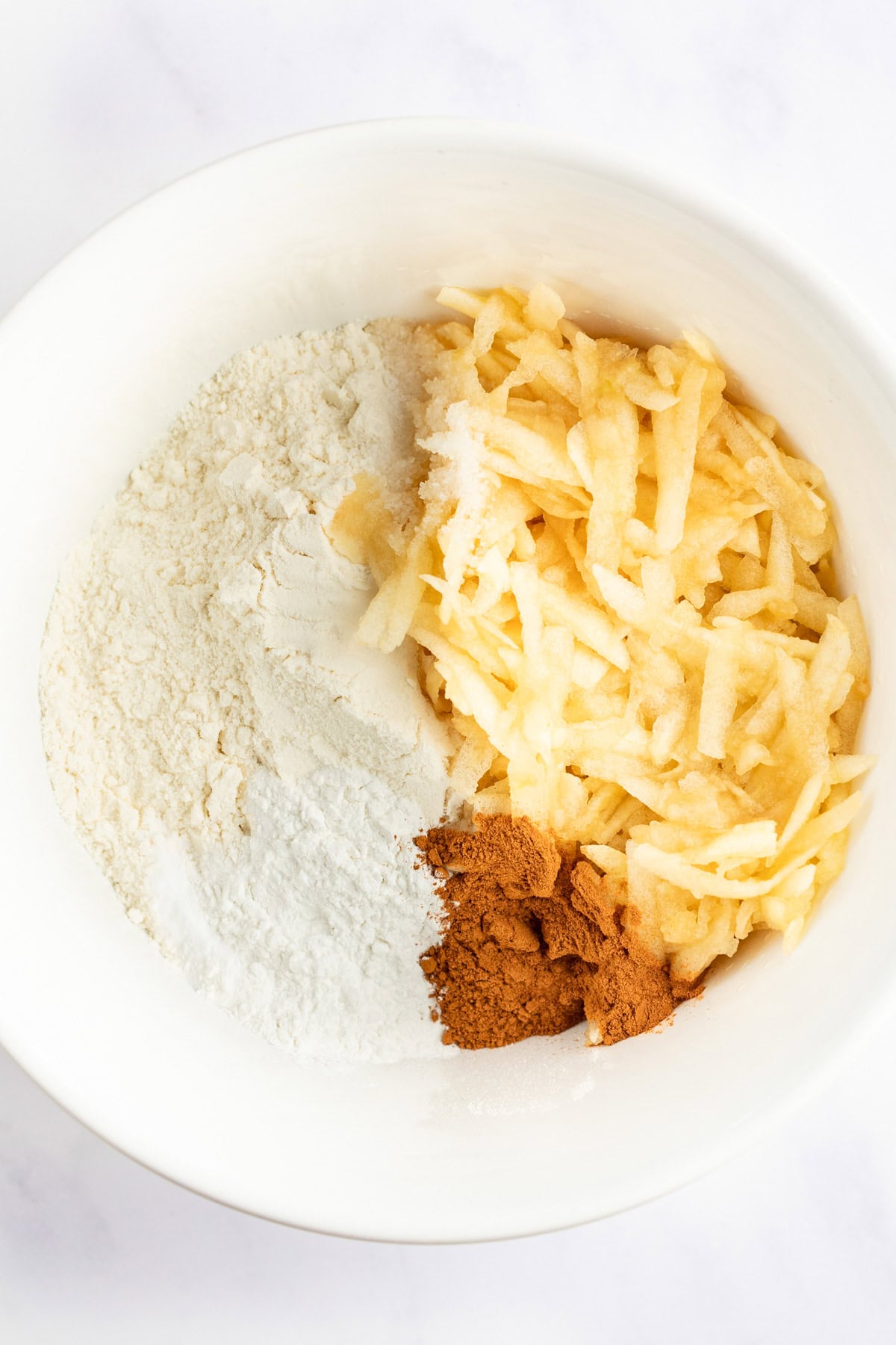 A white mixing bowl containing flour, grated apples, and a portion of ground cinnamon.