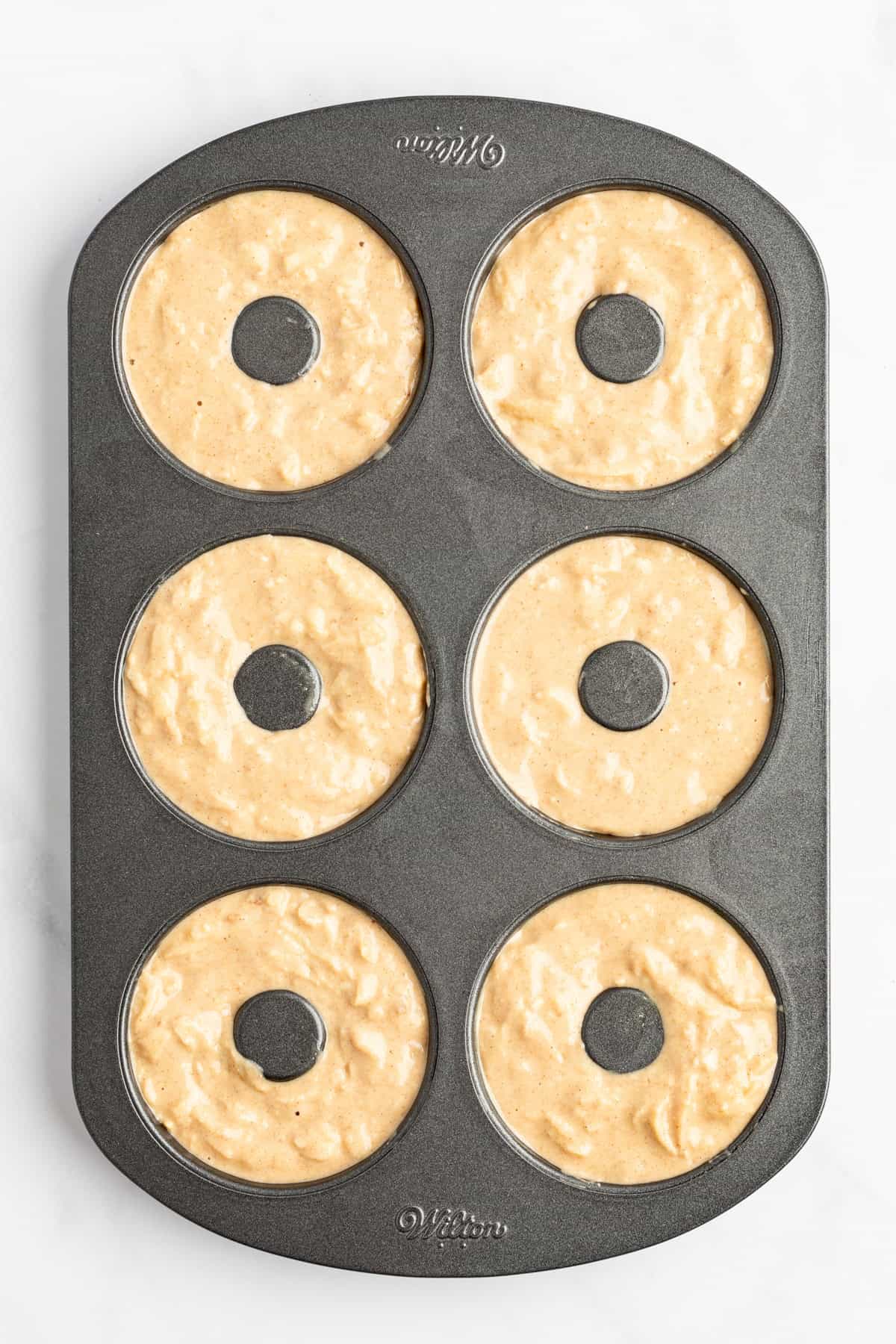 A six-cavity donut baking pan filled with unbaked donut batter, ready to be placed in the oven.