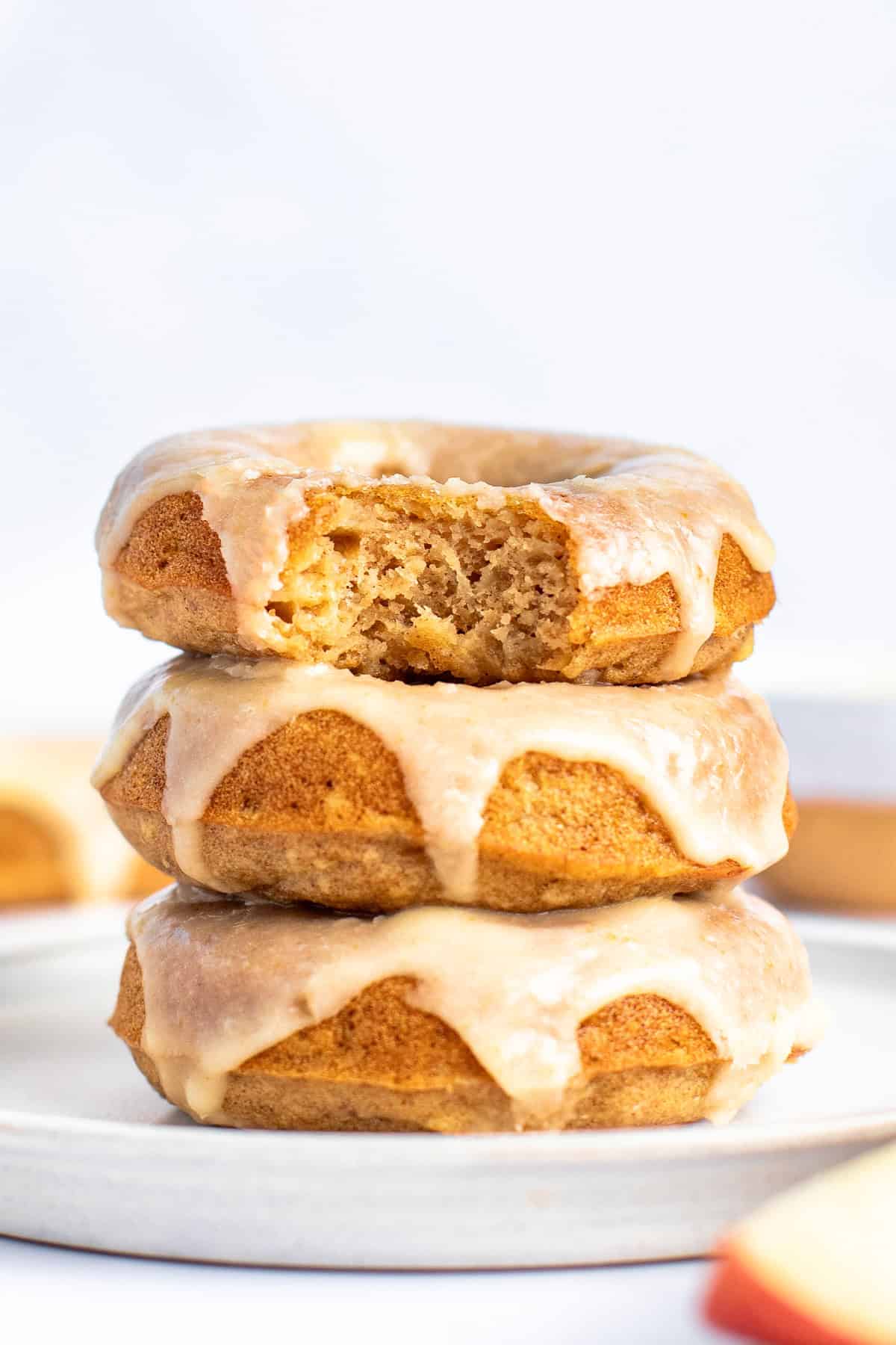 A stack of three glazed donuts on a white plate, with the top donut partially eaten, showing a golden brown interior.