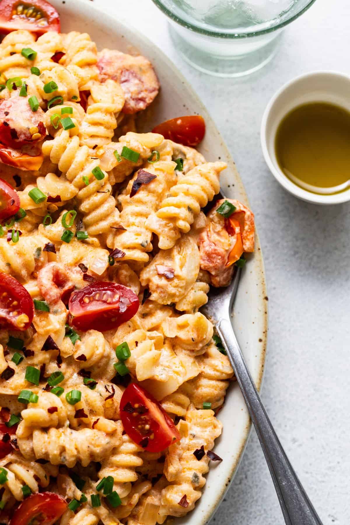 A dish of creamy pasta with sliced sausage, cherry tomatoes, and garnished with chopped green onions, served with a side of olive oil.