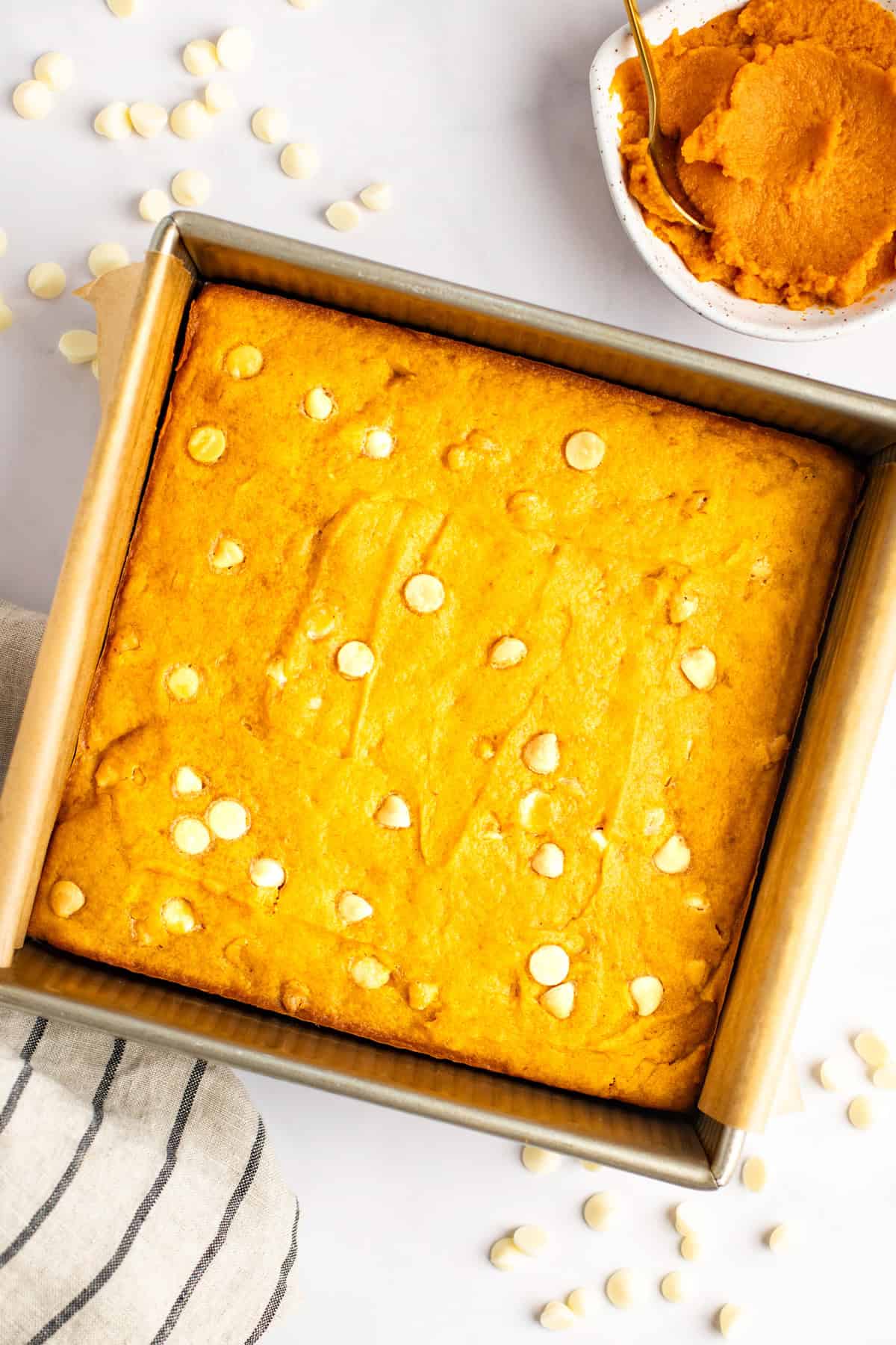 A square pan of orange-colored baked goods with white chocolate chips. A small bowl of pumpkin puree and a spoon are placed beside the pan. A striped cloth and scattered chocolate chips are also visible.