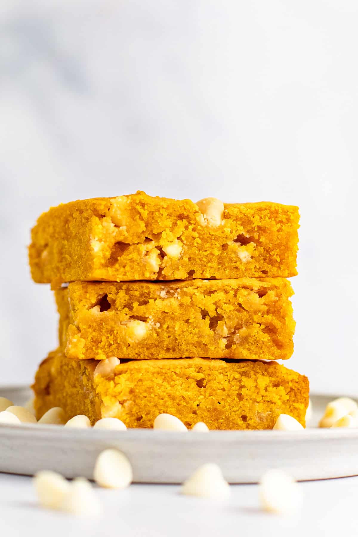 A stack of three pumpkin-flavored blondies with white chocolate chips sits on a grey plate, surrounded by scattered white chocolate chips, against a white background.