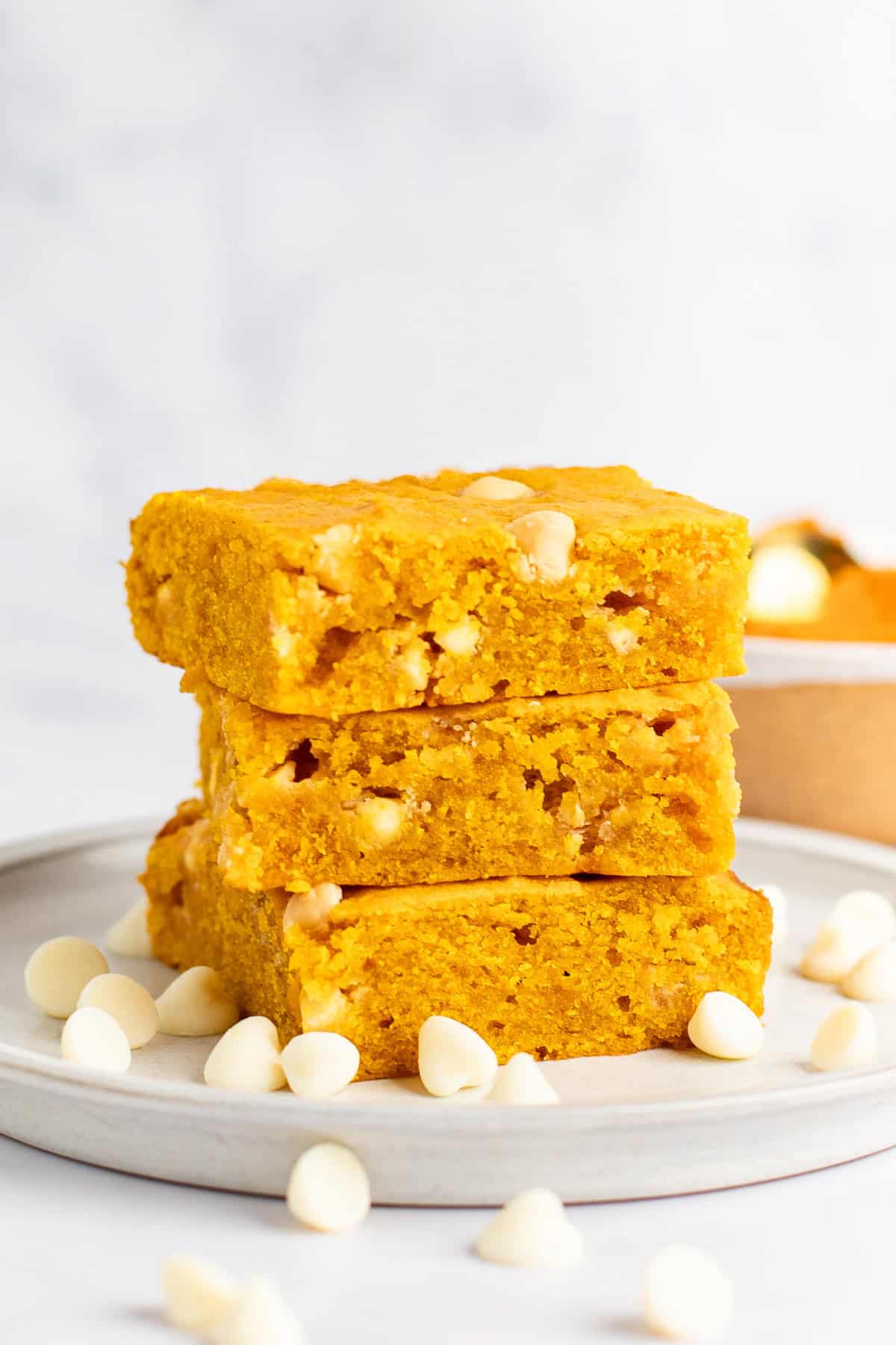 Three slices of golden yellow pumpkin blondies stacked on a plate, surrounded by white chocolate chips.