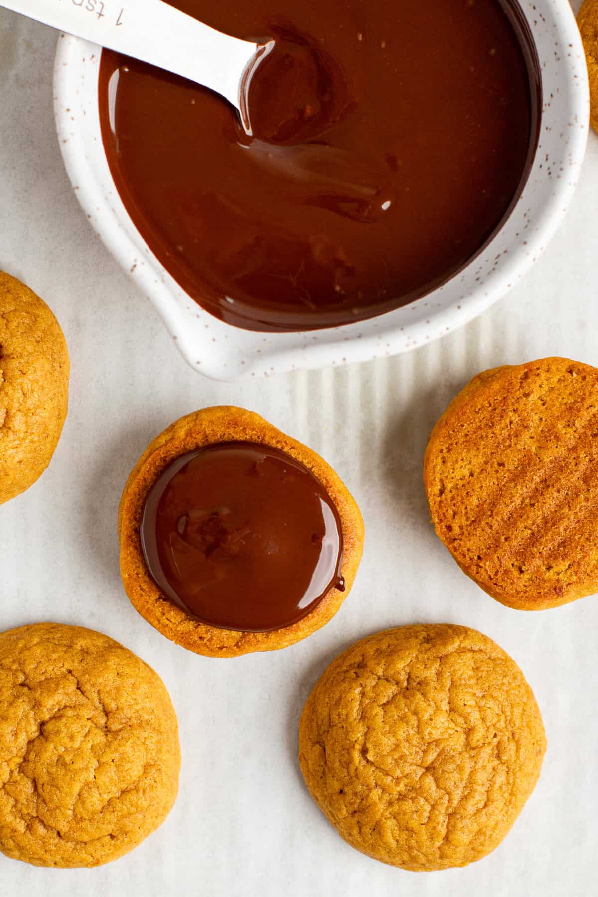 Cookies being dipped in melted chocolate from a white bowl, with additional cookies placed nearby on a white surface.