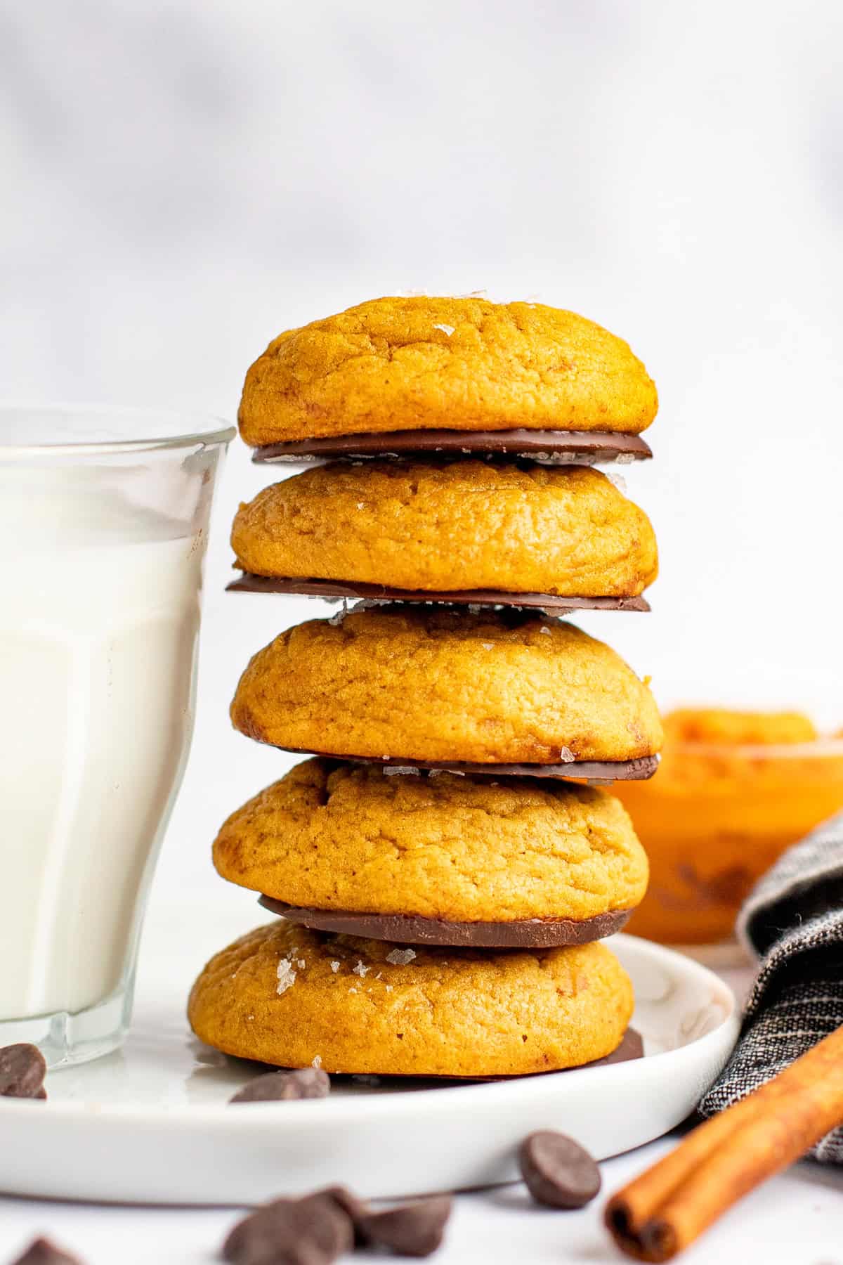 A stack of five pumpkin cookies on a white plate with a glass of milk on the left and a folded cloth napkin on the right, with chocolate chips scattered around.