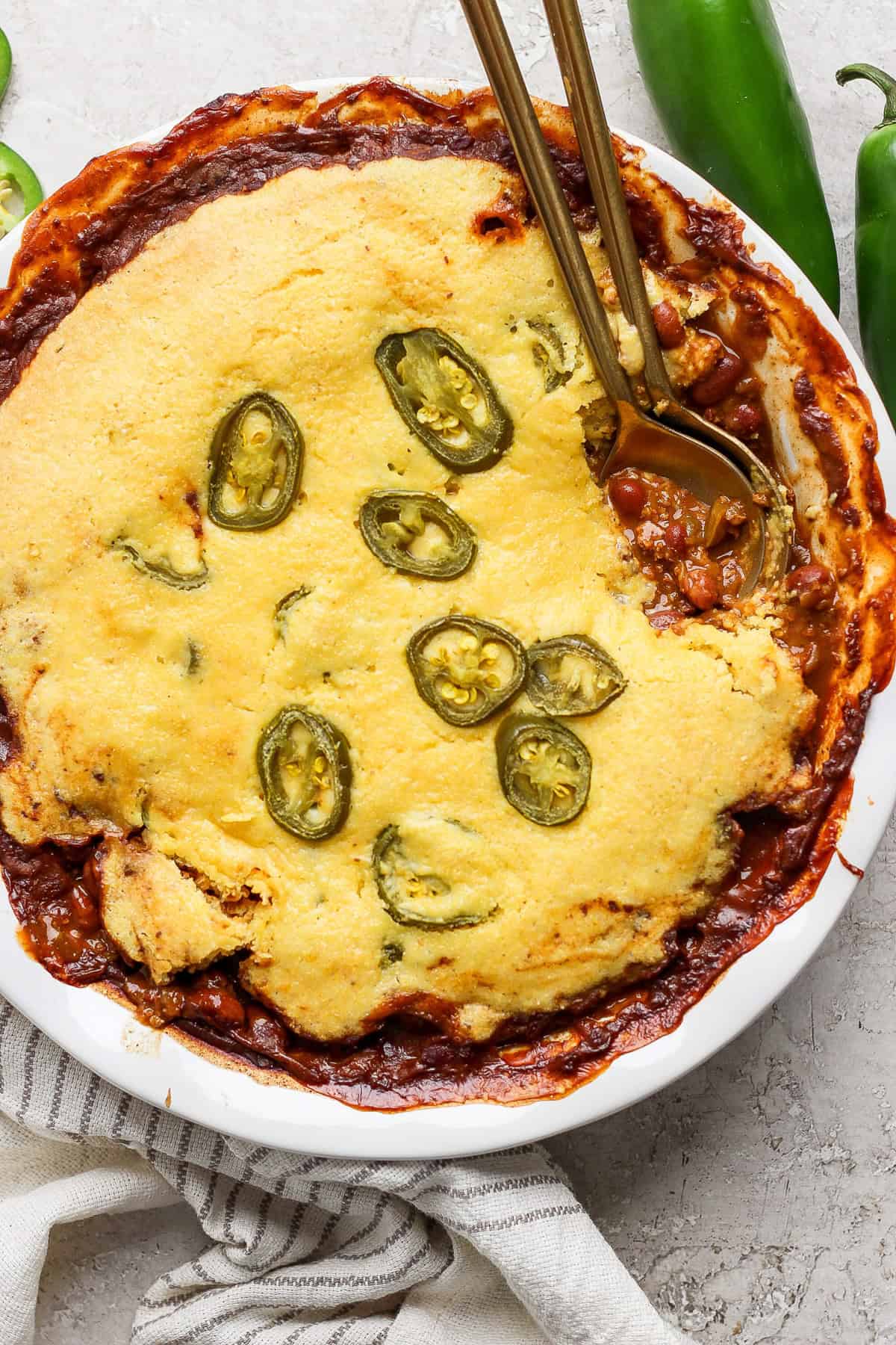 A dish of cornbread chili pie topped with sliced jalapeños, served in a white pie dish, with two gold-colored spoons placed on top, set on a light-colored surface.