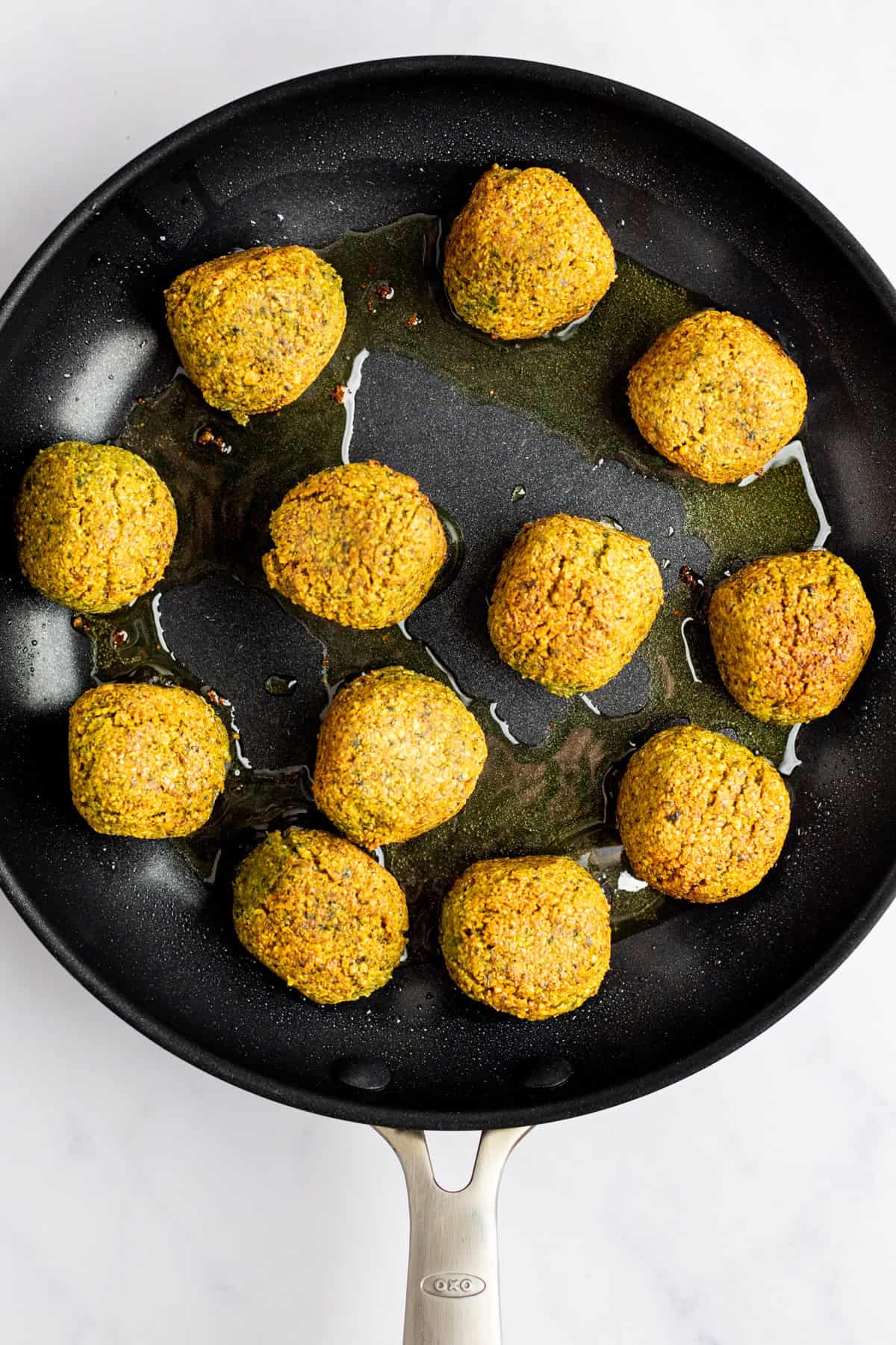 A skillet with twelve golden-brown falafel balls frying in oil on a light-colored surface.