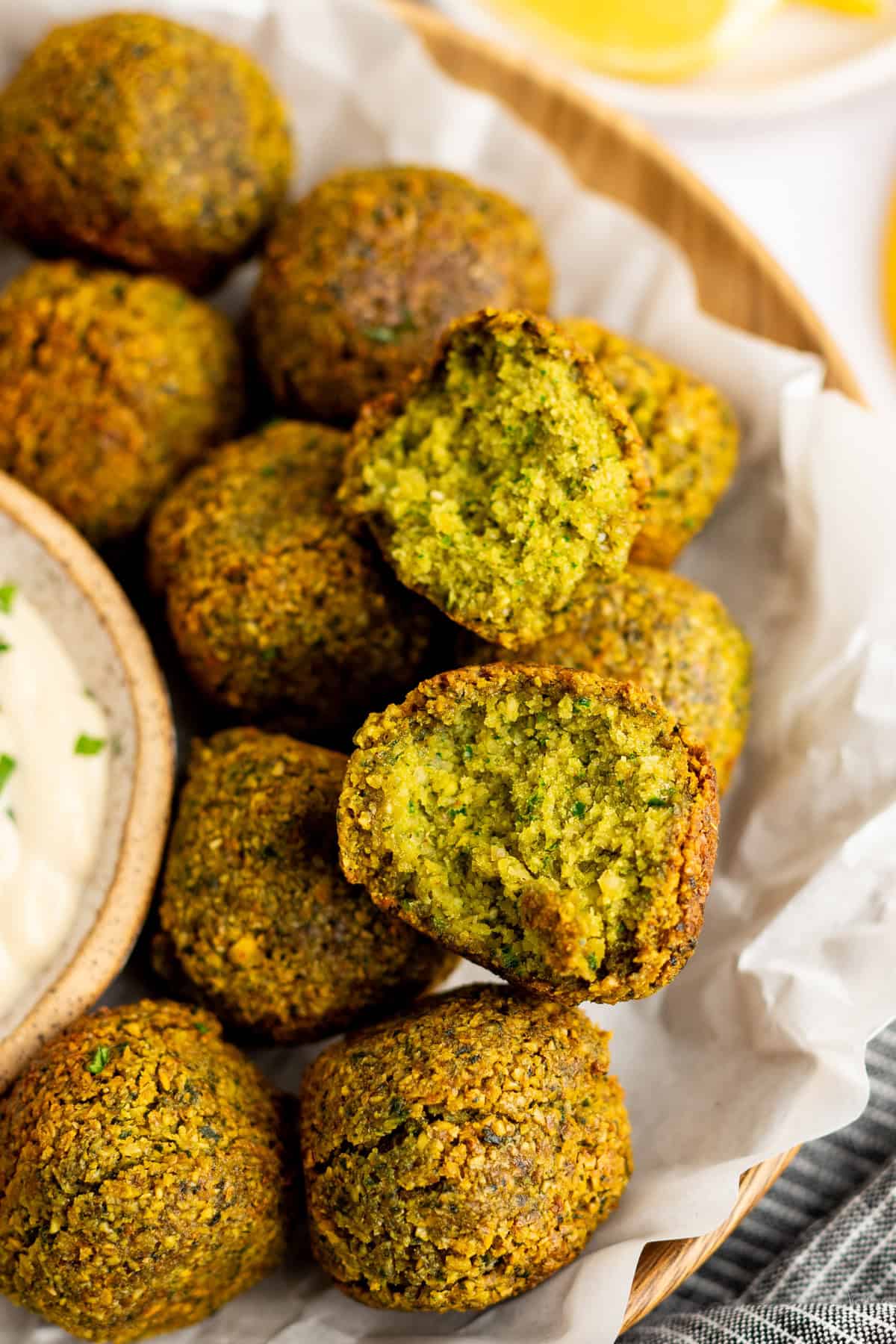 A close-up of a basket of falafel balls, with one broken open to show the green interior. A small bowl of dipping sauce is partially visible next to the falafel.