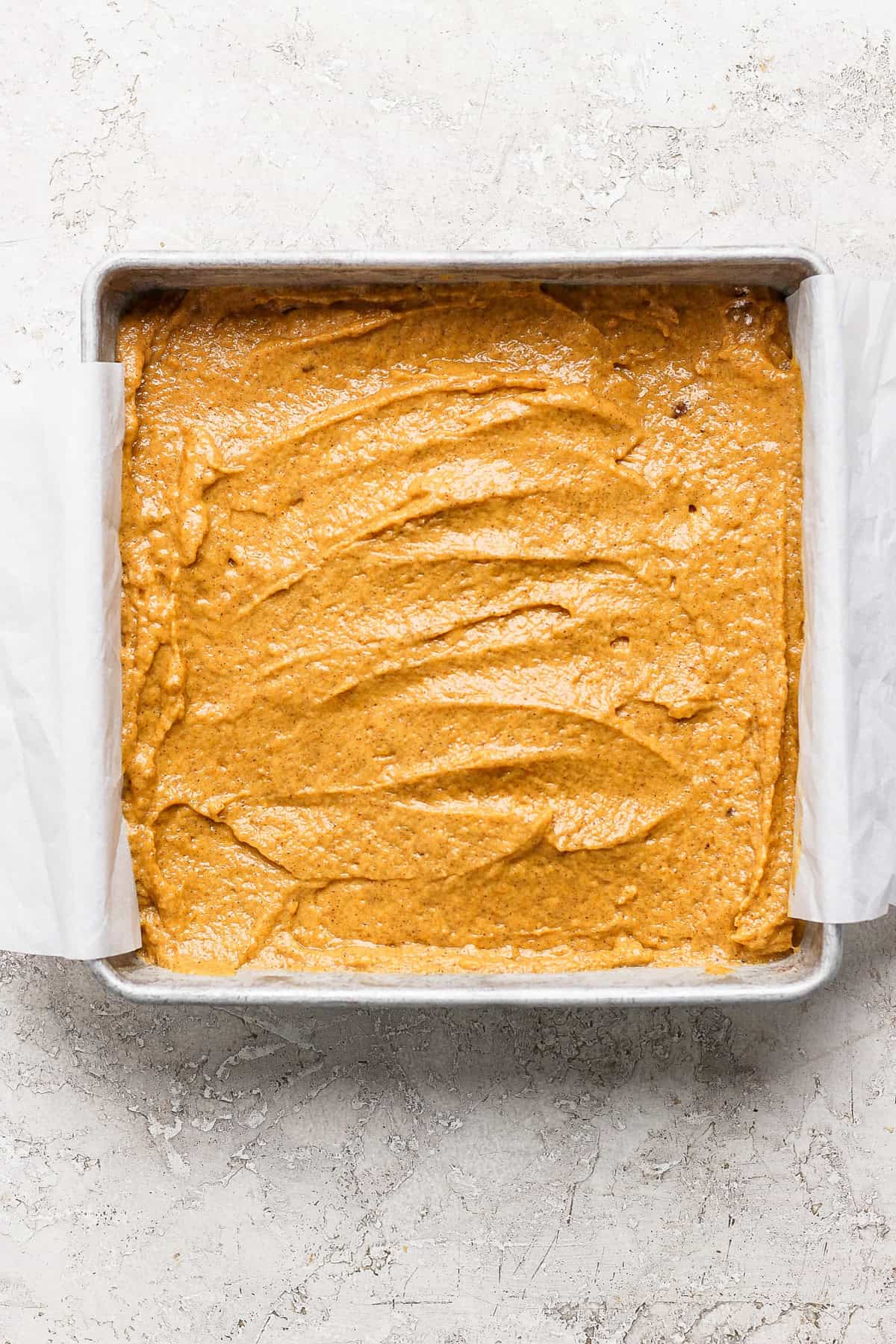 Square baking pan filled with unbaked pumpkin batter on a light textured surface. Edges of the parchment paper extend out from the pan.