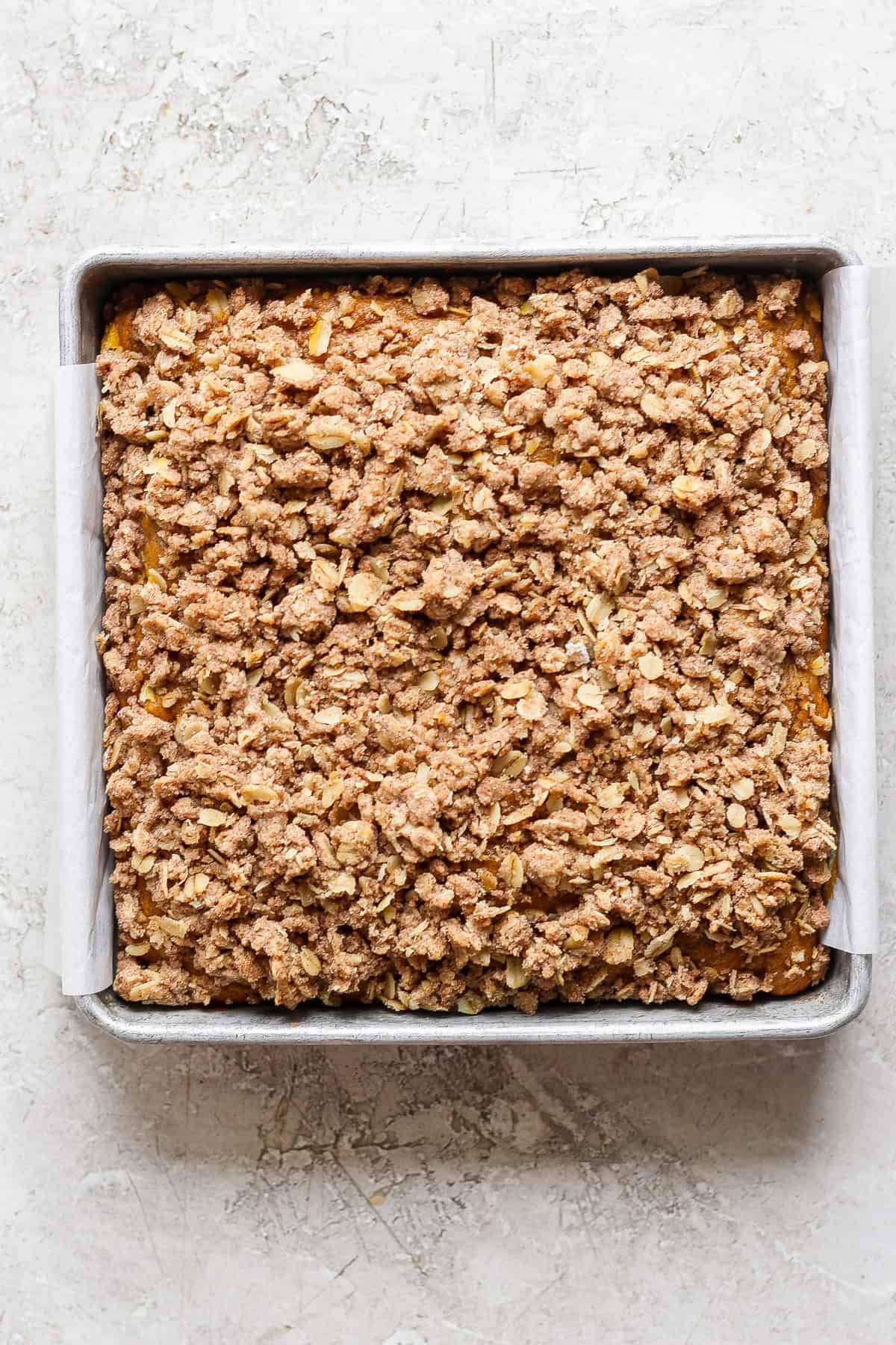 A square metal baking pan filled with a baked oatmeal fruit crumble topped with a crispy, golden-brown oat and cinnamon streusel. The pan is lined with parchment paper.