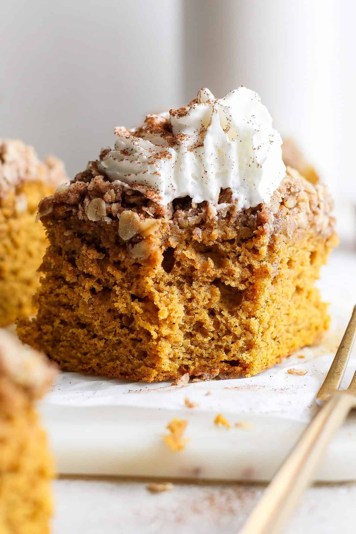 A close-up of a piece of pumpkin cake topped with streusel and whipped cream, placed on a white surface beside a gold fork.