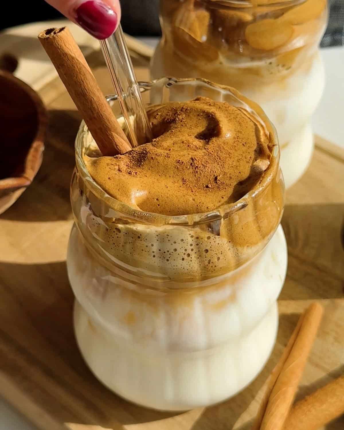 A glass of whipped coffee with a thick foam layer on top, garnished with a cinnamon stick, sits on a wooden board next to a second similar drink.