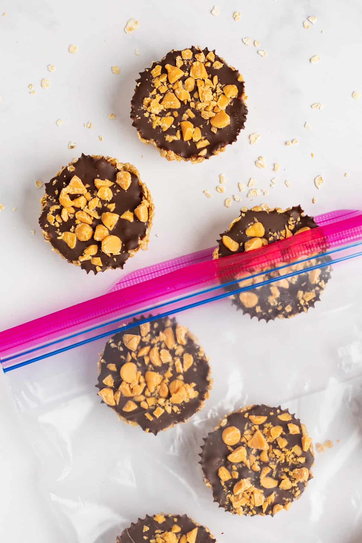 Five chocolate peanut butter cups topped with crushed peanuts, with three inside a partially open ziplock bag and two outside, on a white surface with scattered peanut pieces.