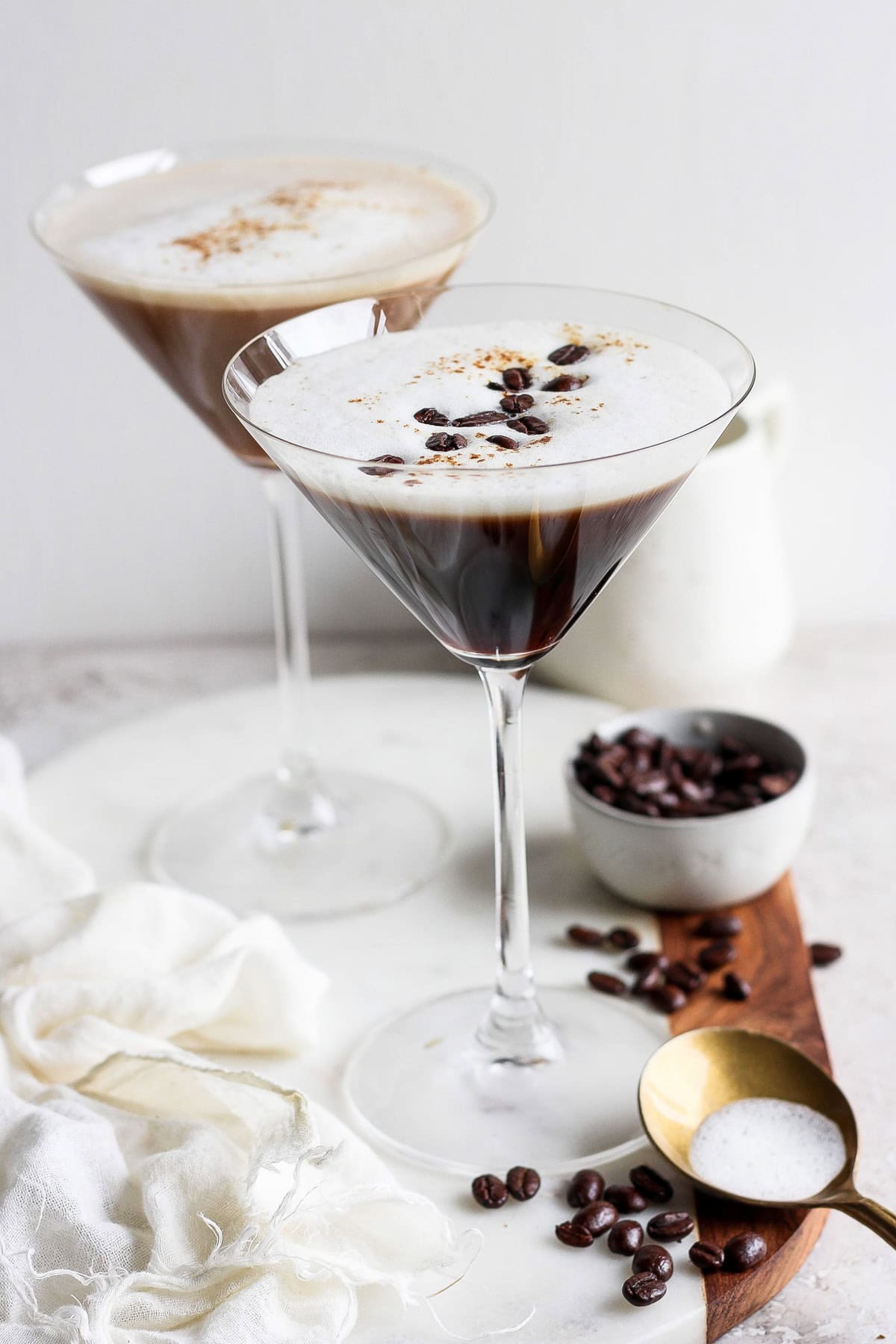 Two espresso martinis in martini glasses, garnished with coffee beans, on a wooden board with a small bowl of coffee beans and a brass spoon nearby. A white cloth is draped on the left.