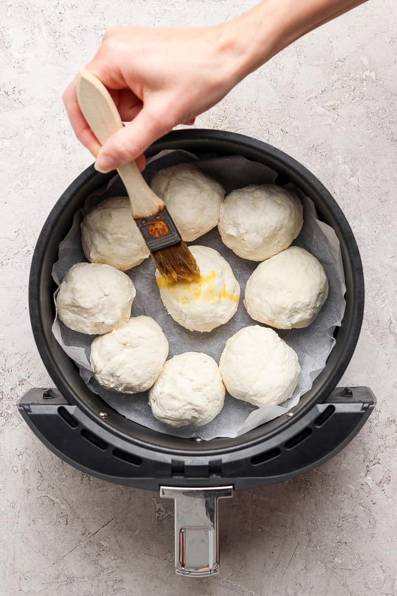 A hand brushes egg wash on dough balls arranged in an air fryer basket lined with parchment paper.