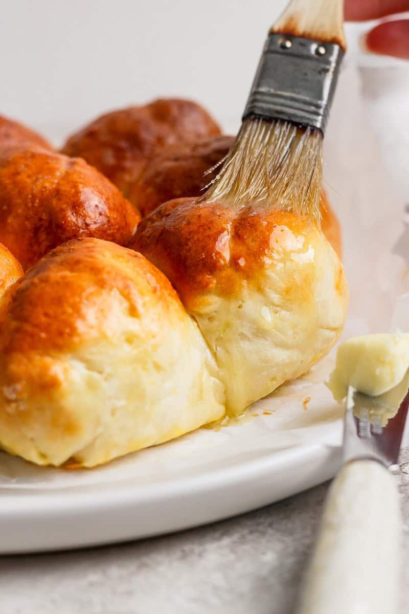 A pastry brush applies butter to freshly baked golden-brown bread rolls on a white plate.