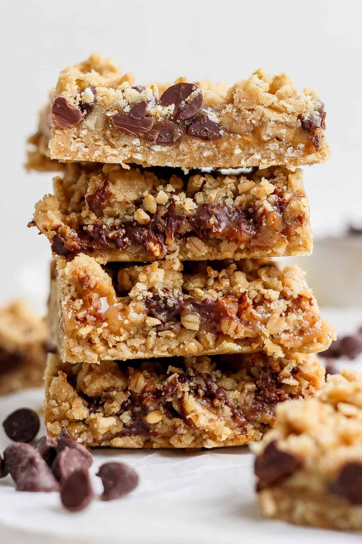 A stack of four chocolate chip oatmeal bars placed on a white surface, with scattered chocolate chips around them.