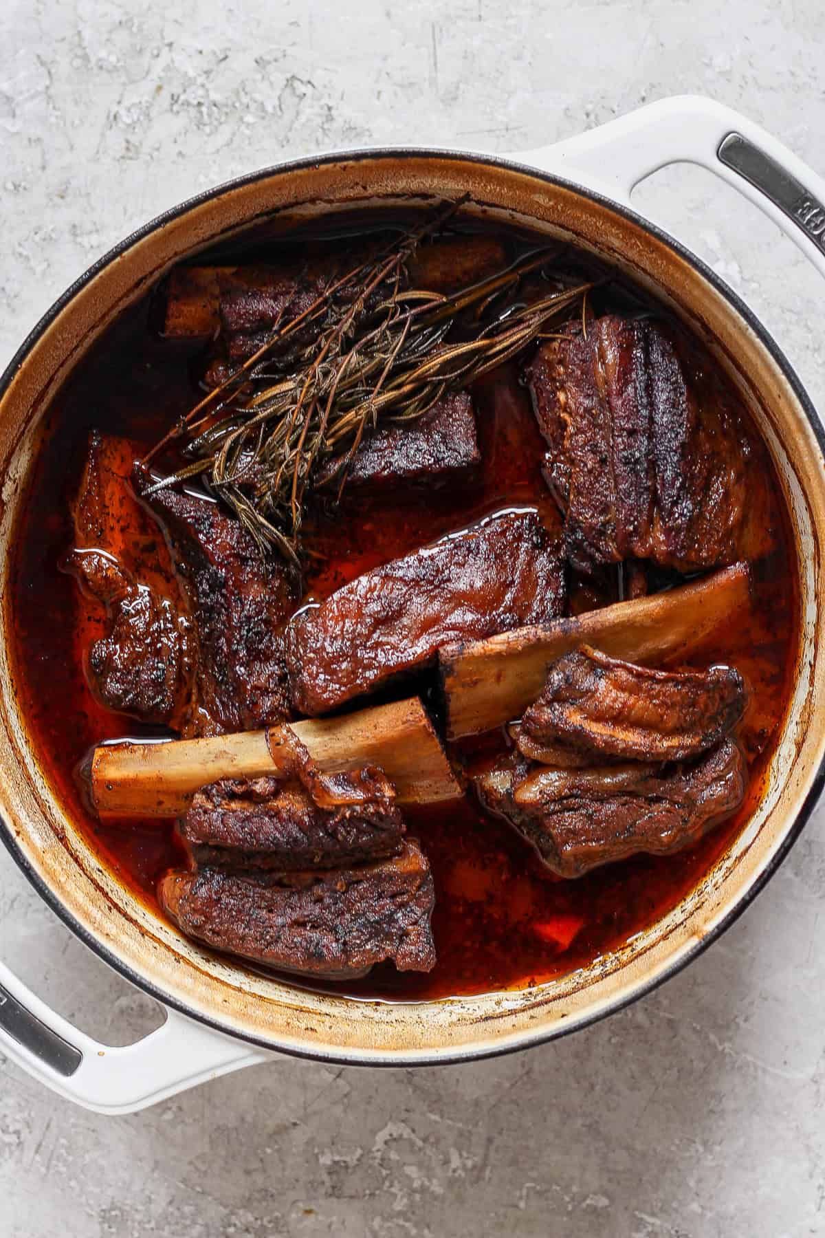 A large pot filled with braised short ribs in a rich, dark sauce, garnished with herbs on a light gray surface.