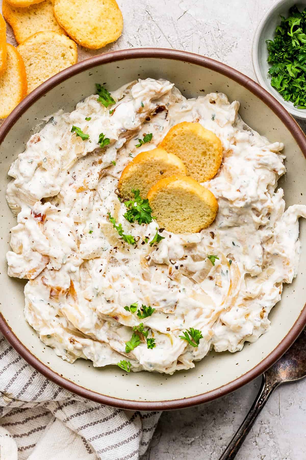 A bowl of creamy dip topped with herbs and three round crackers, placed on a textured surface with more crackers and a side of chopped parsley.