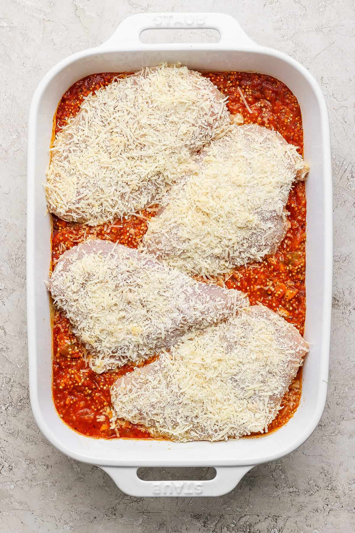 Four uncooked chicken breasts covered in grated cheese are placed on top of a layer of tomato sauce in a white baking dish.