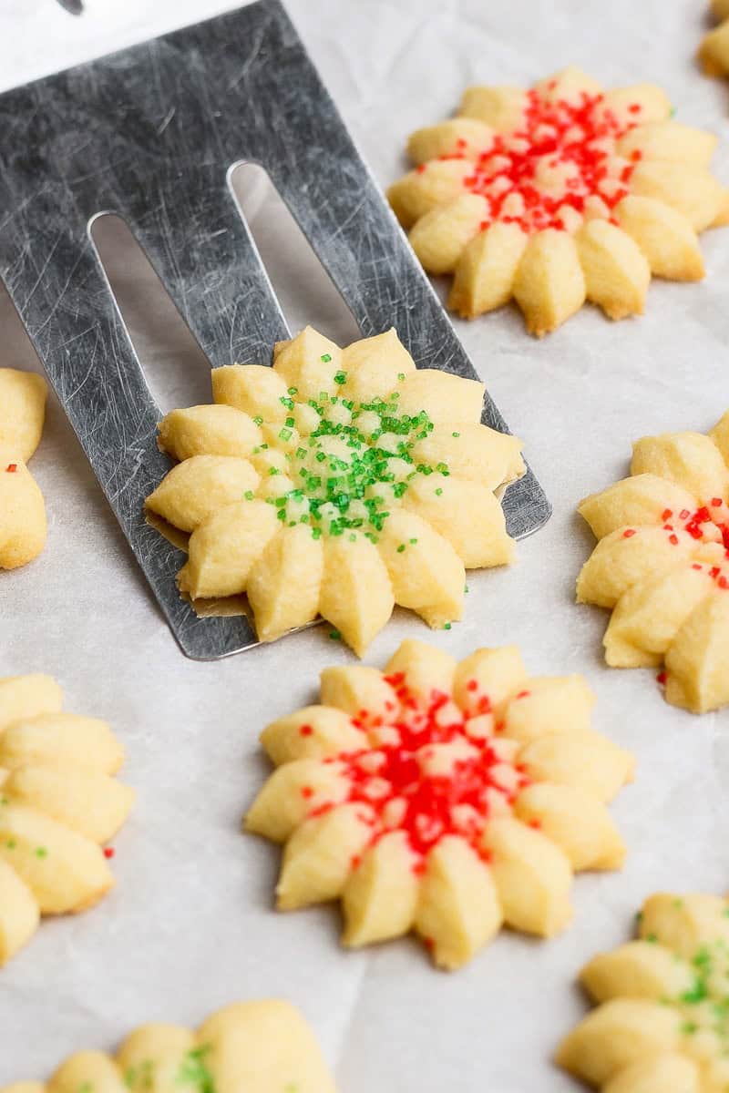 Butter cookies decorated with red and green sprinkles, arranged on a baking sheet. A spatula lifts one cookie.