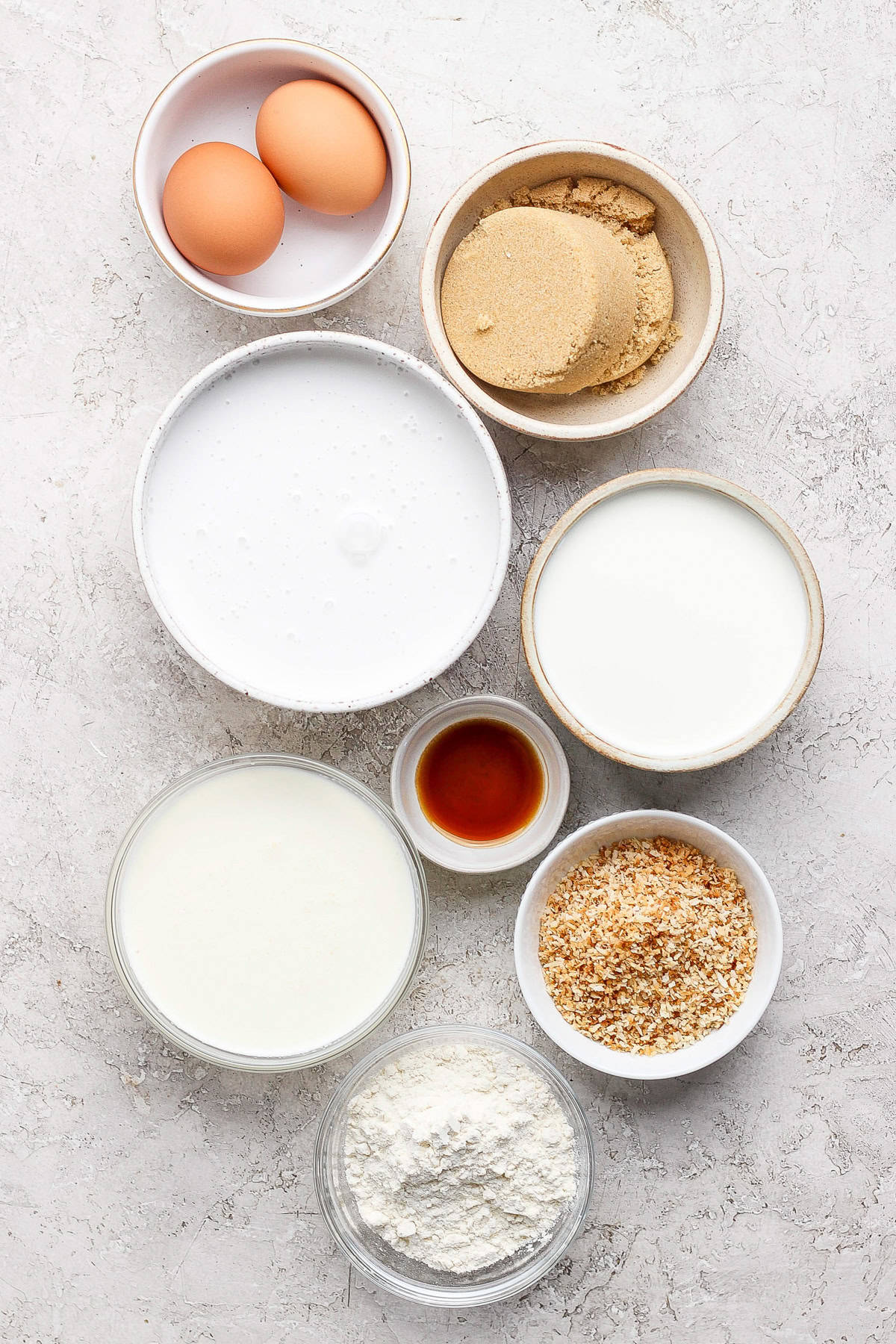 Assorted baking ingredients including eggs, brown sugar, flour, vanilla extract, coconut flakes, and milk, arranged on a light surface.