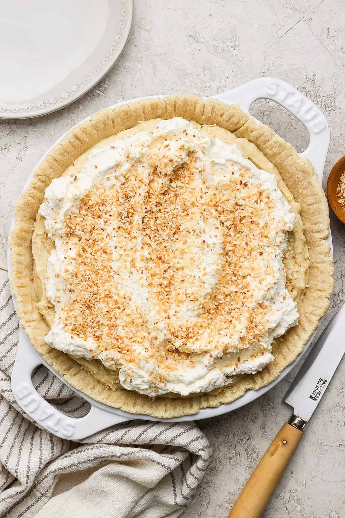 A pie topped with whipped cream and toasted coconut flakes, on a light-colored surface with a knife and striped cloth nearby.