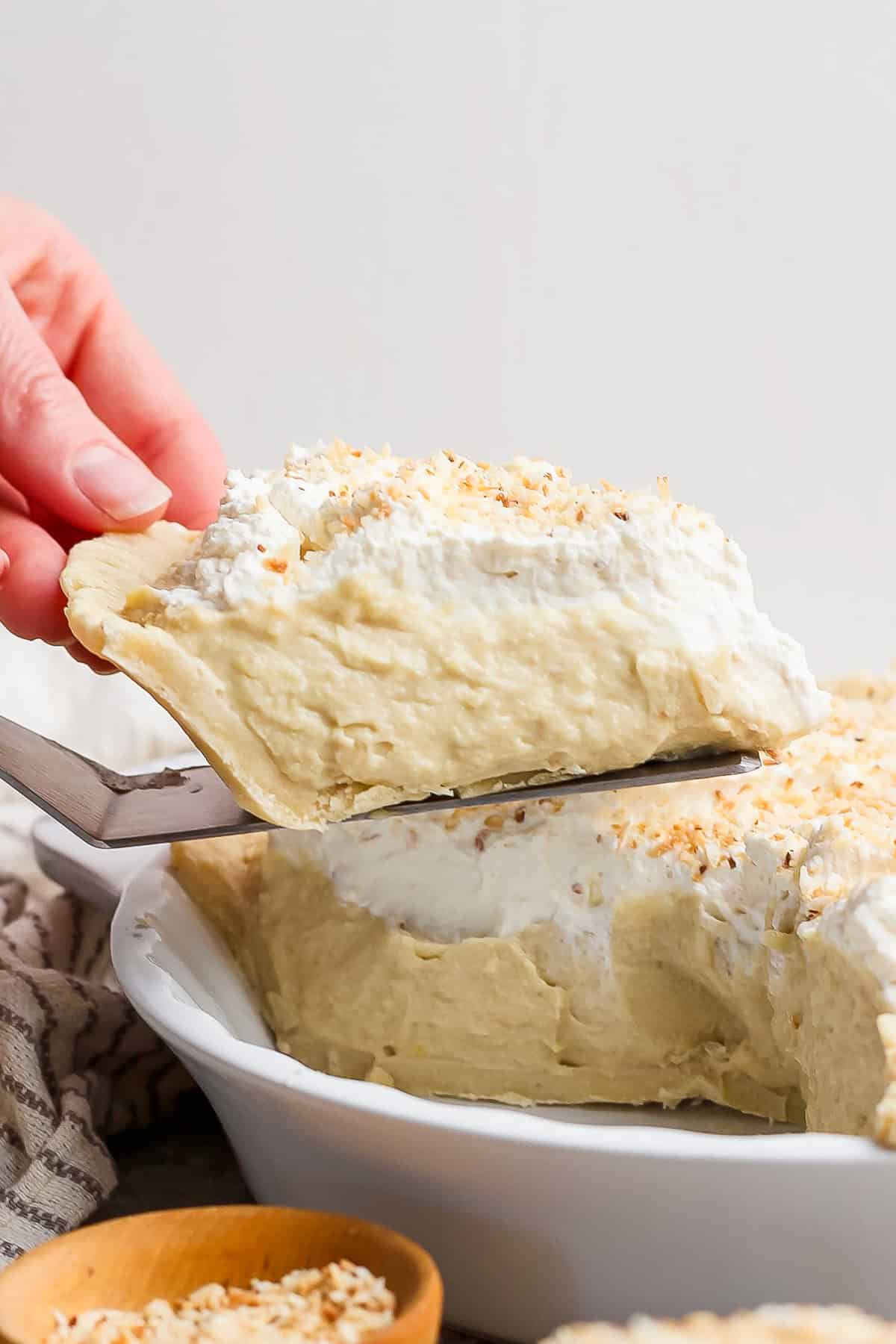 A hand serves a slice of creamy pie topped with whipped cream and sprinkled with coconut from a white dish.